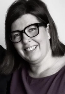 Black and white photo of a woman with glasses, smiling, wearing a dark top.