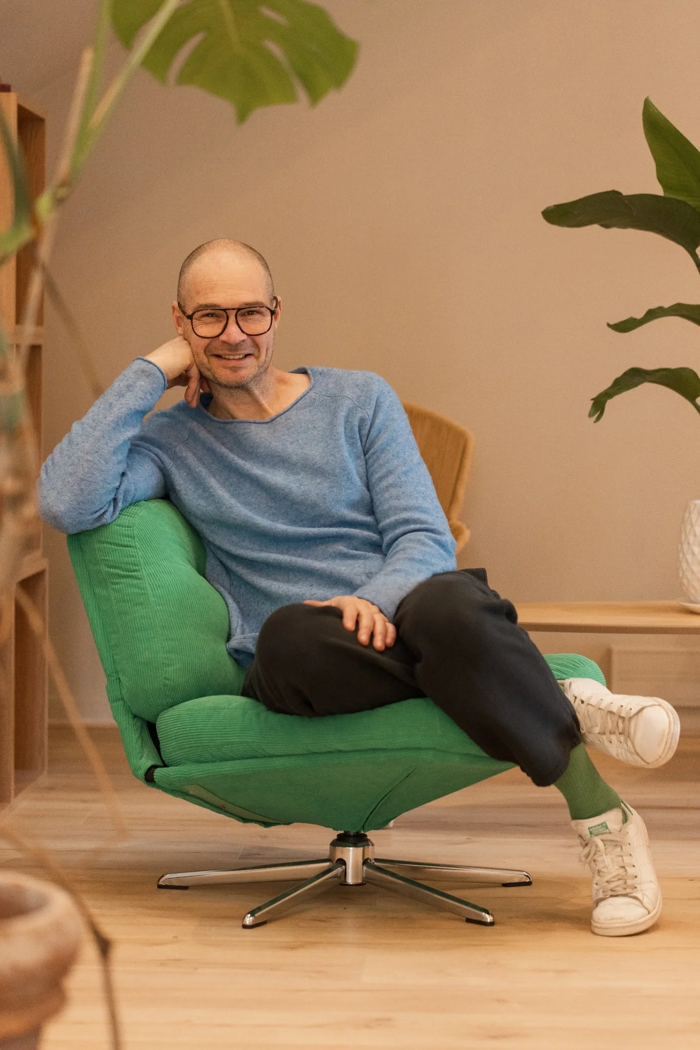 Homme assis confortablement dans un fauteuil vert, portant des lunettes, un sweat bleu et des baskets blanches, dans une pièce avec des plantes et du mobilier en bois.