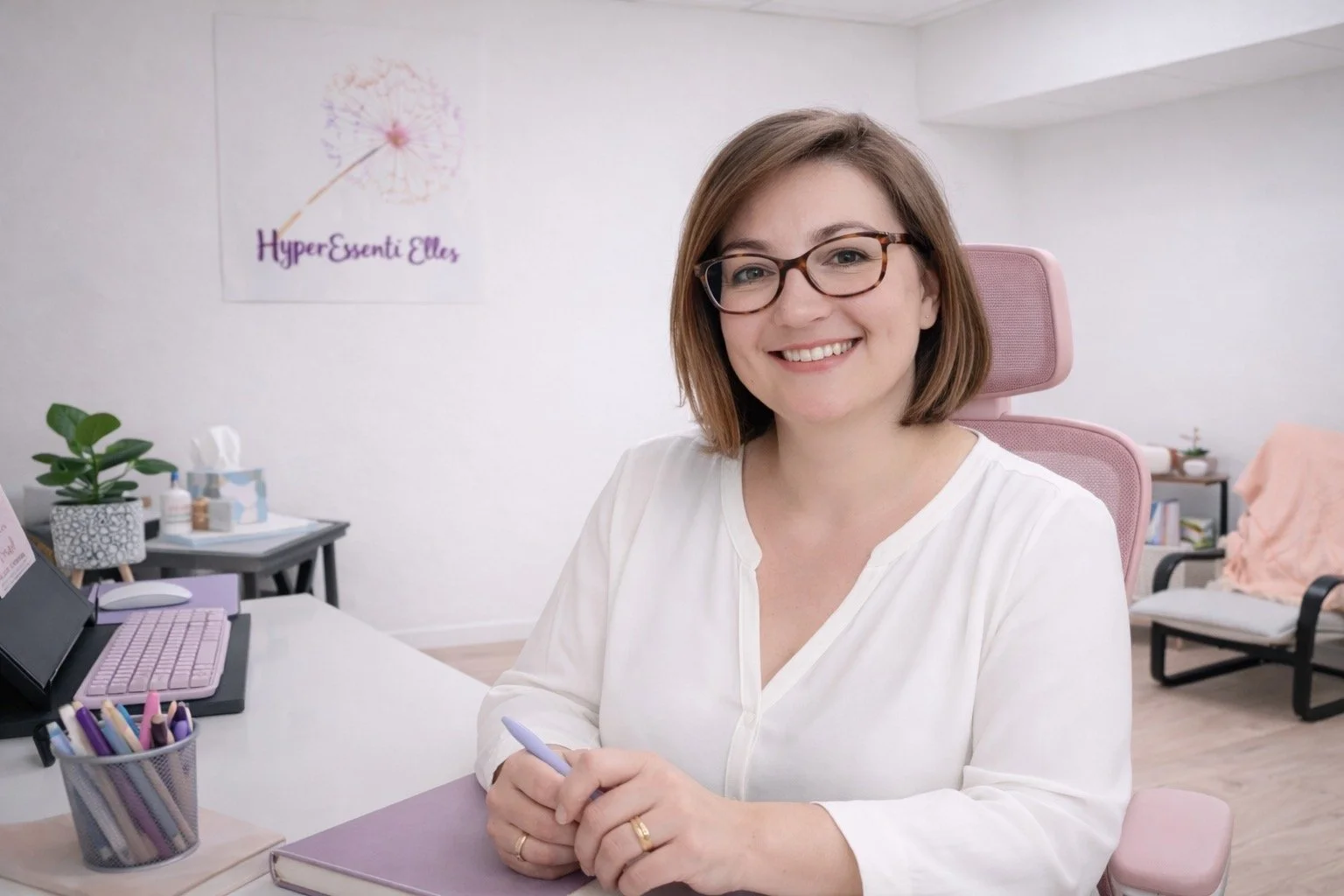 Une femme souriante portant des lunettes, assise à un bureau dans un espace de travail clair et organisé, avec un ordinateur portable, une plante verte, et un tableau sur le mur