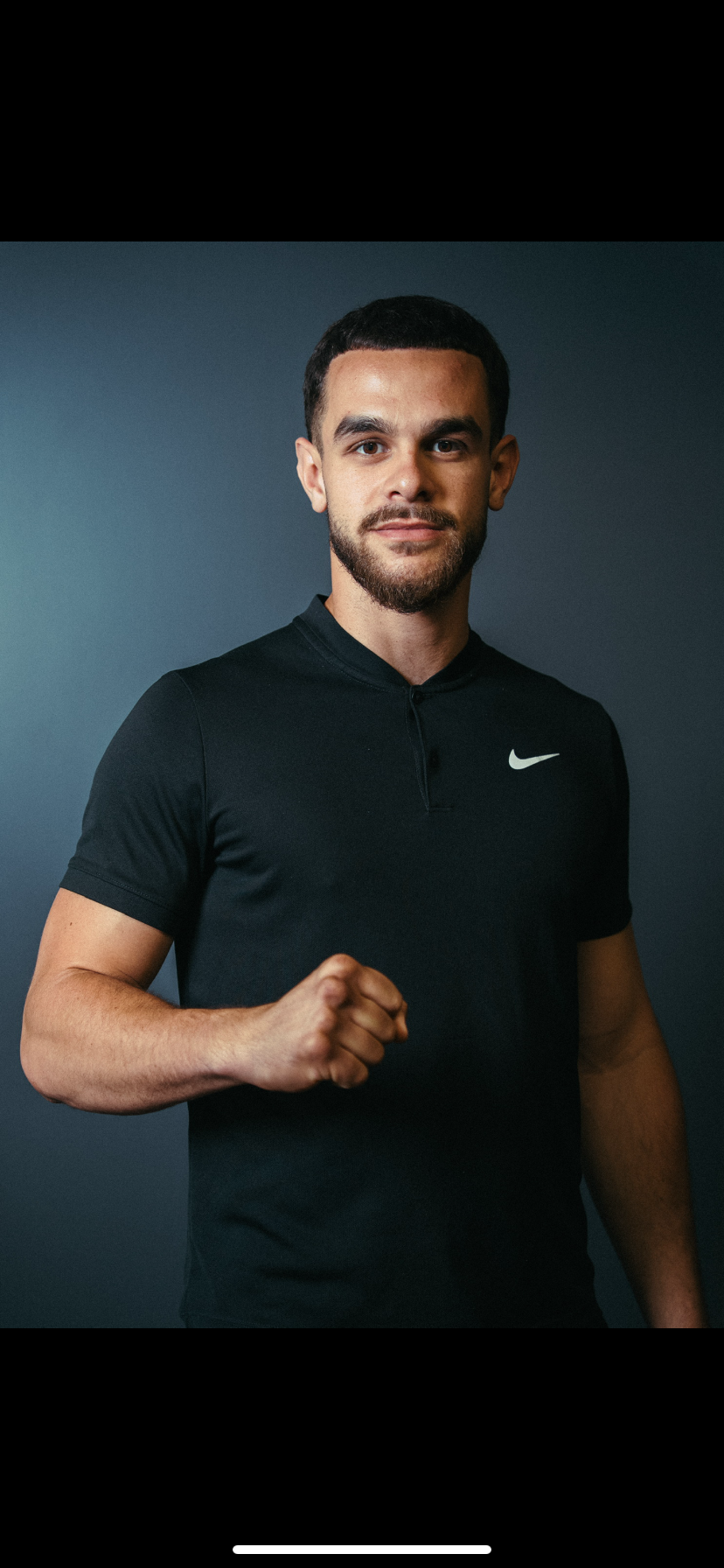 Homme en t-shirt noir Nike, faisant un geste de pouce levé, contre un fond gris foncé.