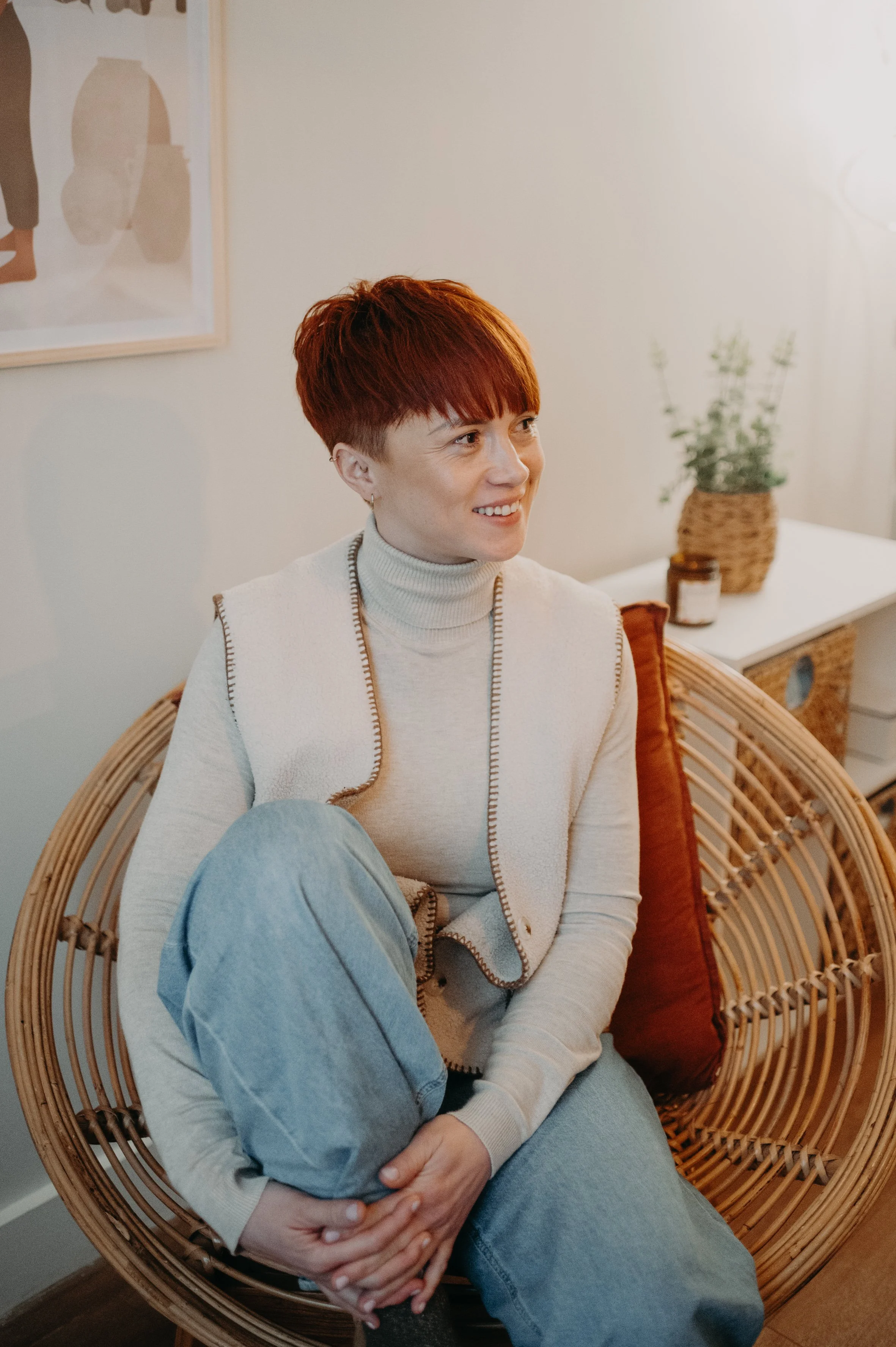 Une femme rousse avec une coupe courte, portant un pull beige avec des détails bruns, assise dans un fauteuil en rotin, souriant, avec un coussin marron derrière elle, dans une pièce avec une plante en pot et un tableau abstrait au mur.