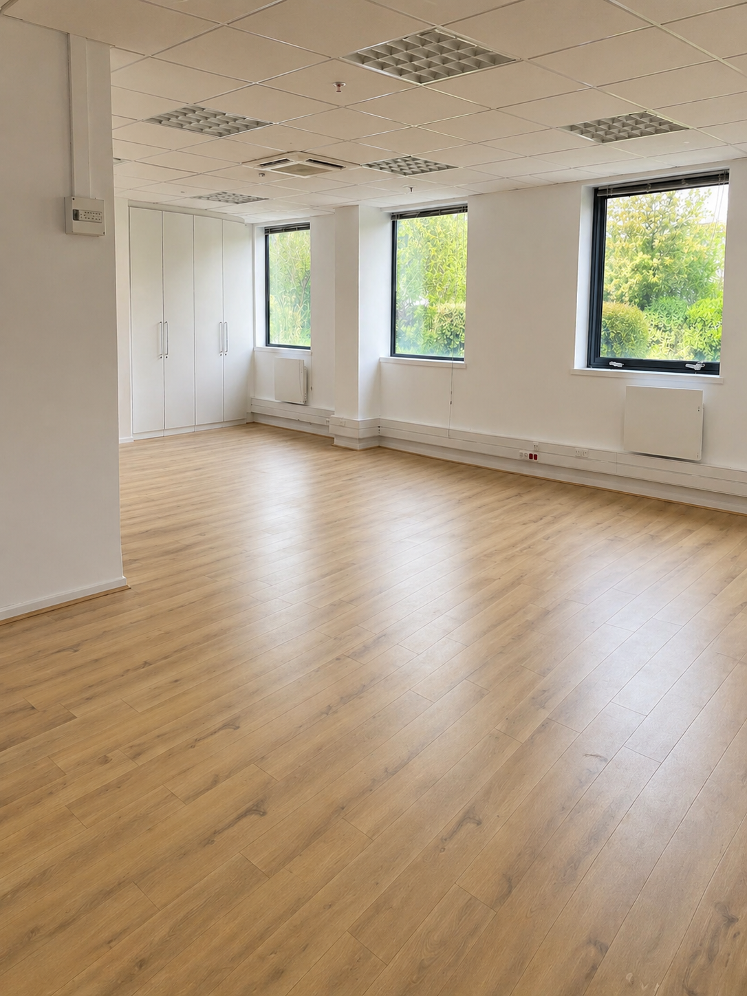 Salle de réunion vide avec fenêtres donnant sur un espace vert, sol en bois clair, murs blancs, et plafonds avec des tuiles suspendues.