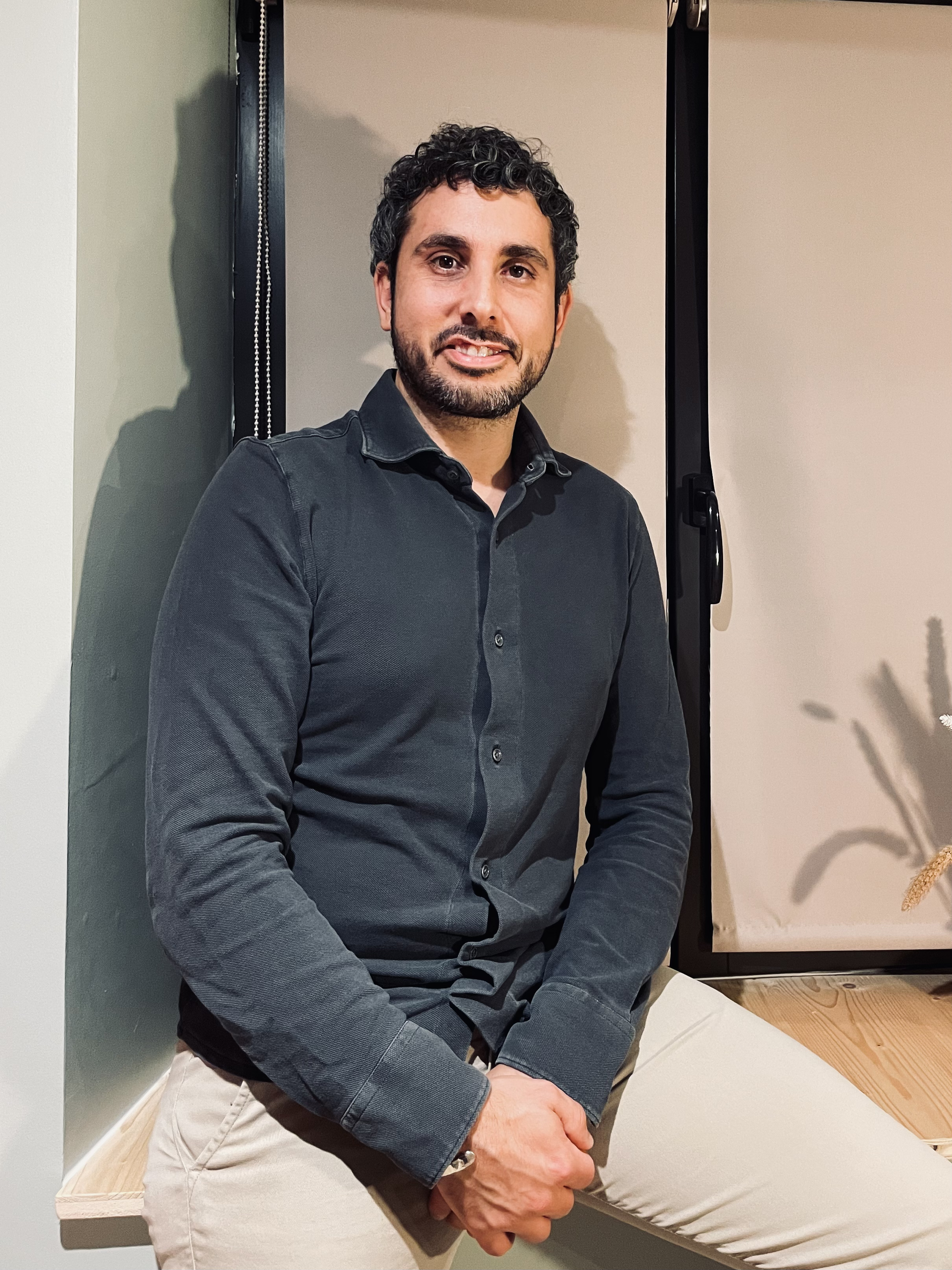 Homme assis sur un banc, portant une chemise noire et un pantalon beige, dans une pièce au décor minimaliste.