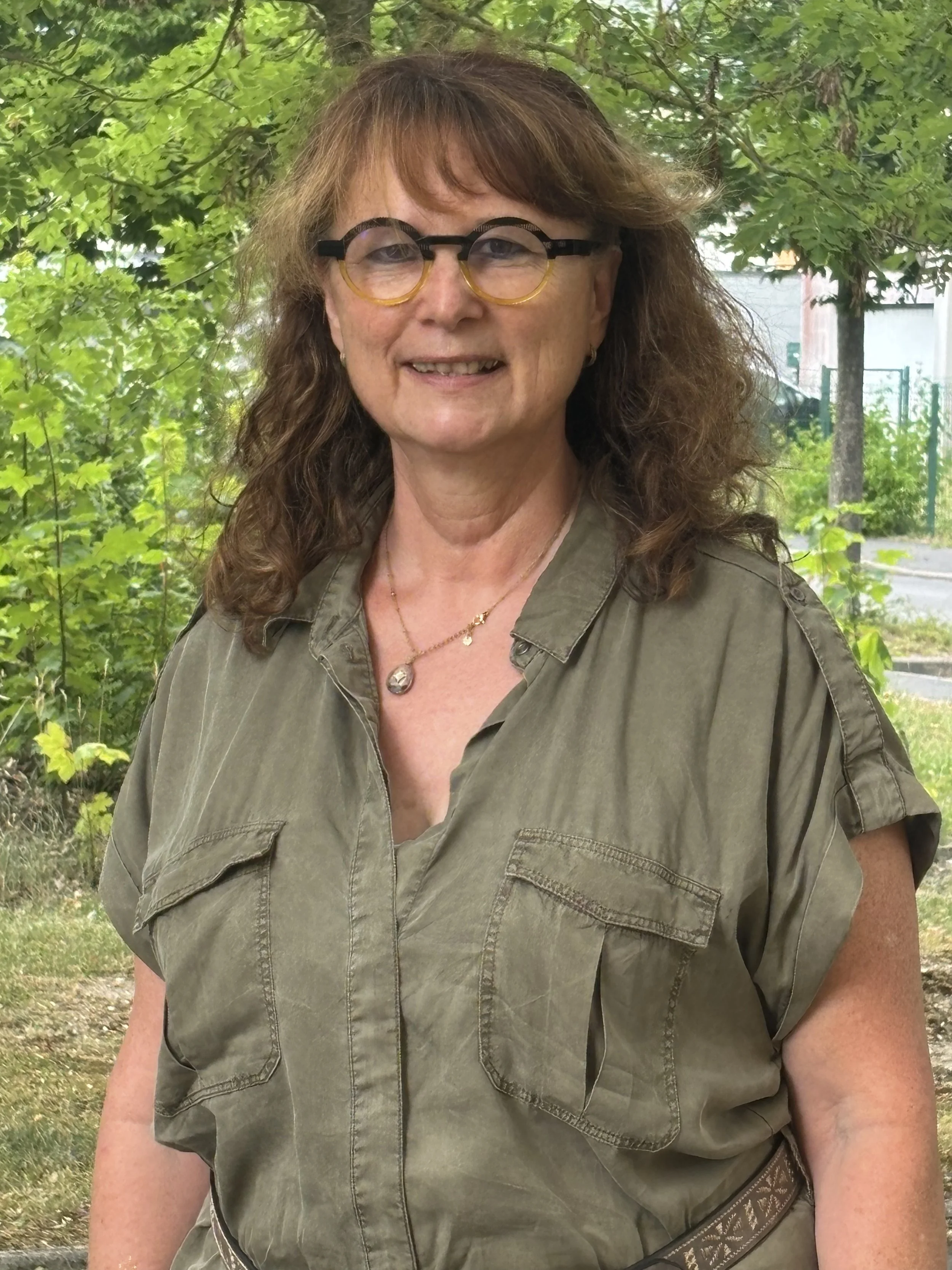 Une femme avec des cheveux bouclés, portant des lunettes rondes et un chemisier vert, se tenant devant des arbres et des buissons dans un environnement extérieur.