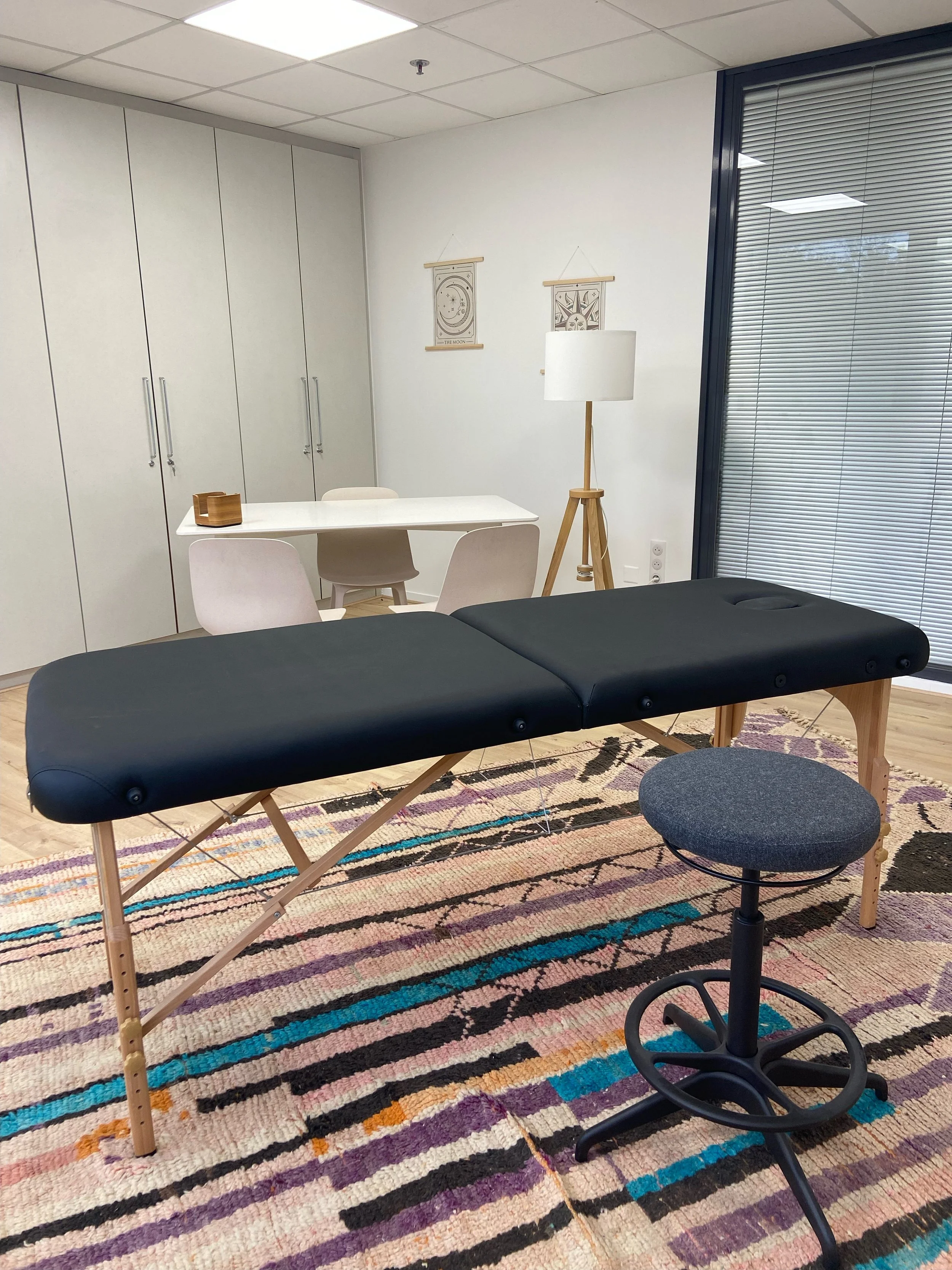 Une salle de consultation ou de massage avec une table noire, une chaise grise, un fauteuil roulant noir, une lampe sur un trépied en bois, deux dessins muraux, un meuble de rangement blanc et une grande fenêtre avec des stores vénitiens.