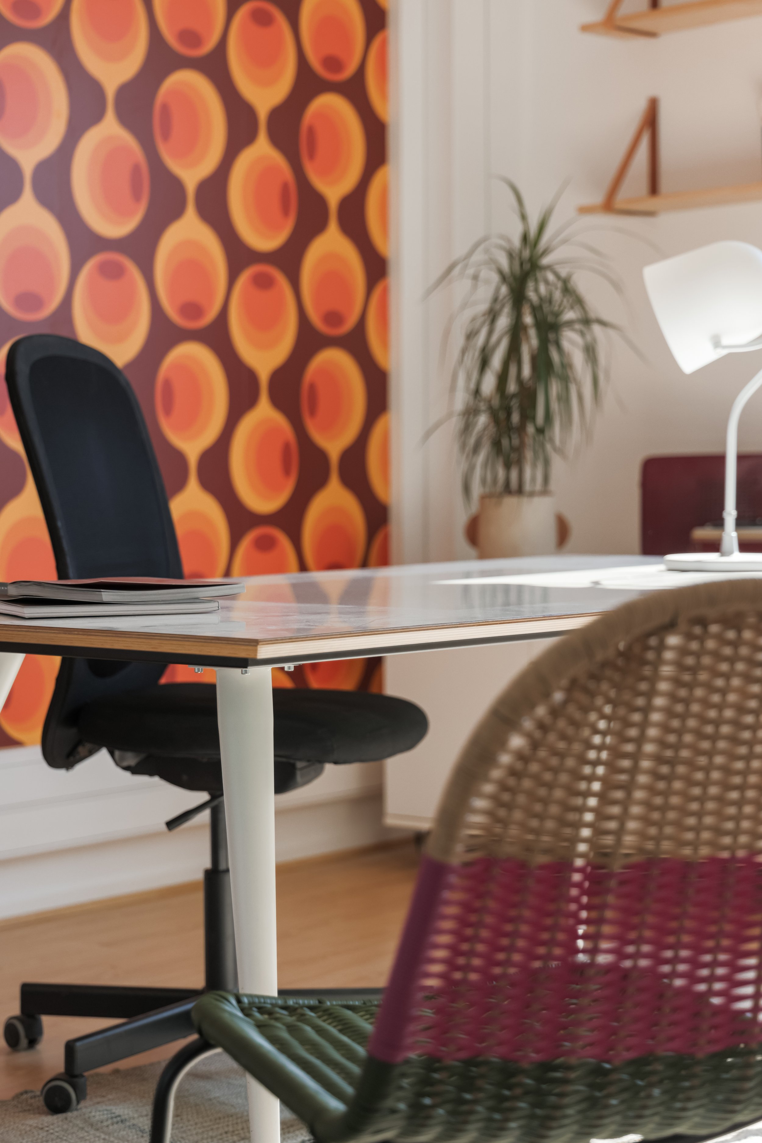 Une salle de réunion ou de coworking avec une table en bois, une chaise de bureau noire, une chaise en rotin avec assise rose, une plante verte en pot, des étagères en bois et une lampe blanche à côté d'un mur blanc et d'un mur décoré avec un papier peint à motifs oranges et marron.