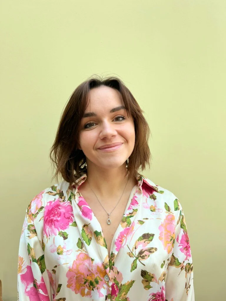 Jeune femme souriante avec des cheveux bruns, portant une chemise à fleurs, devant un fond vert clair.