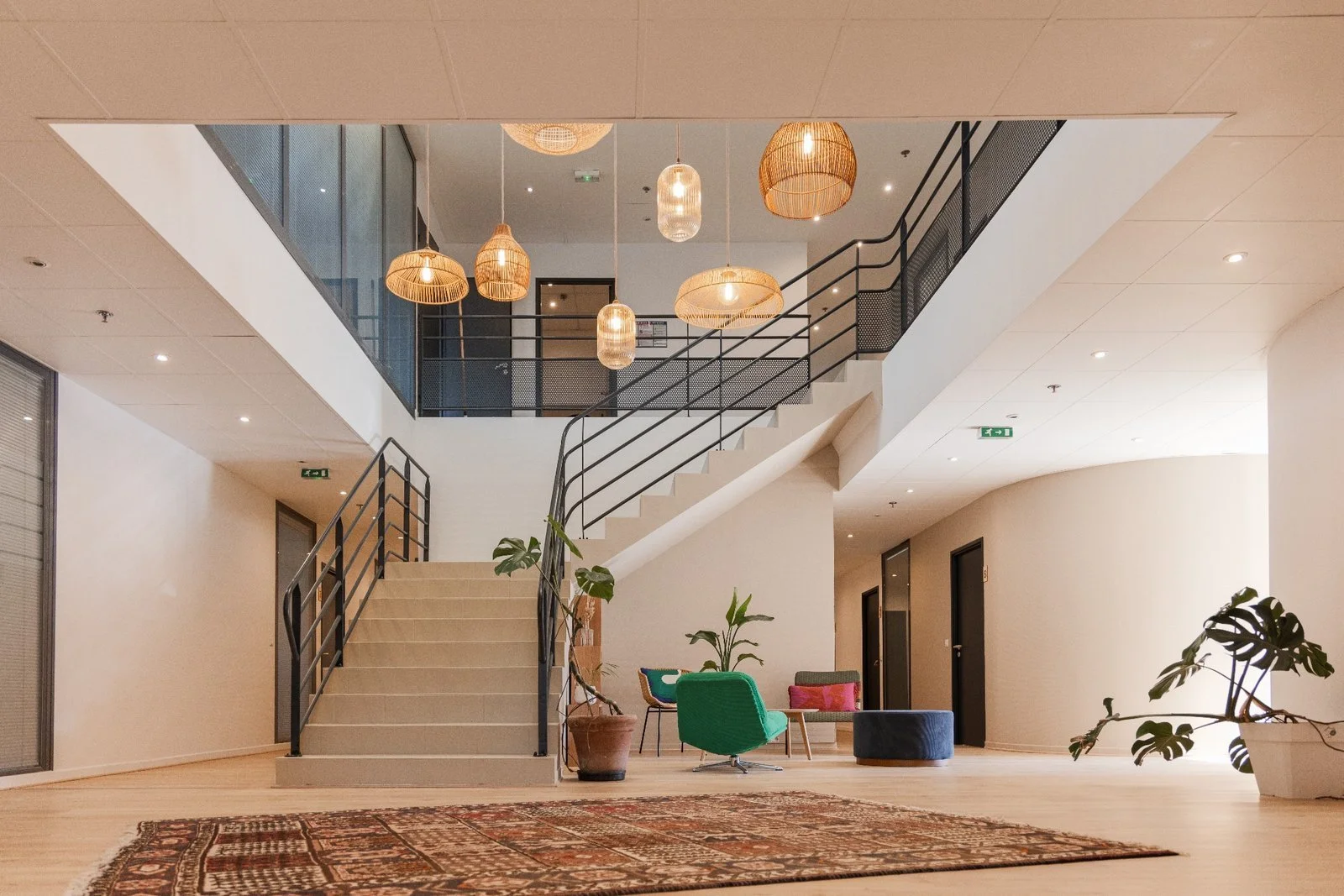 Intérieur moderne avec un escalier central, mobilier contemporain, plantes d'intérieur, lampes suspendues en osier et sol en bois, ambiance chaleureuse.