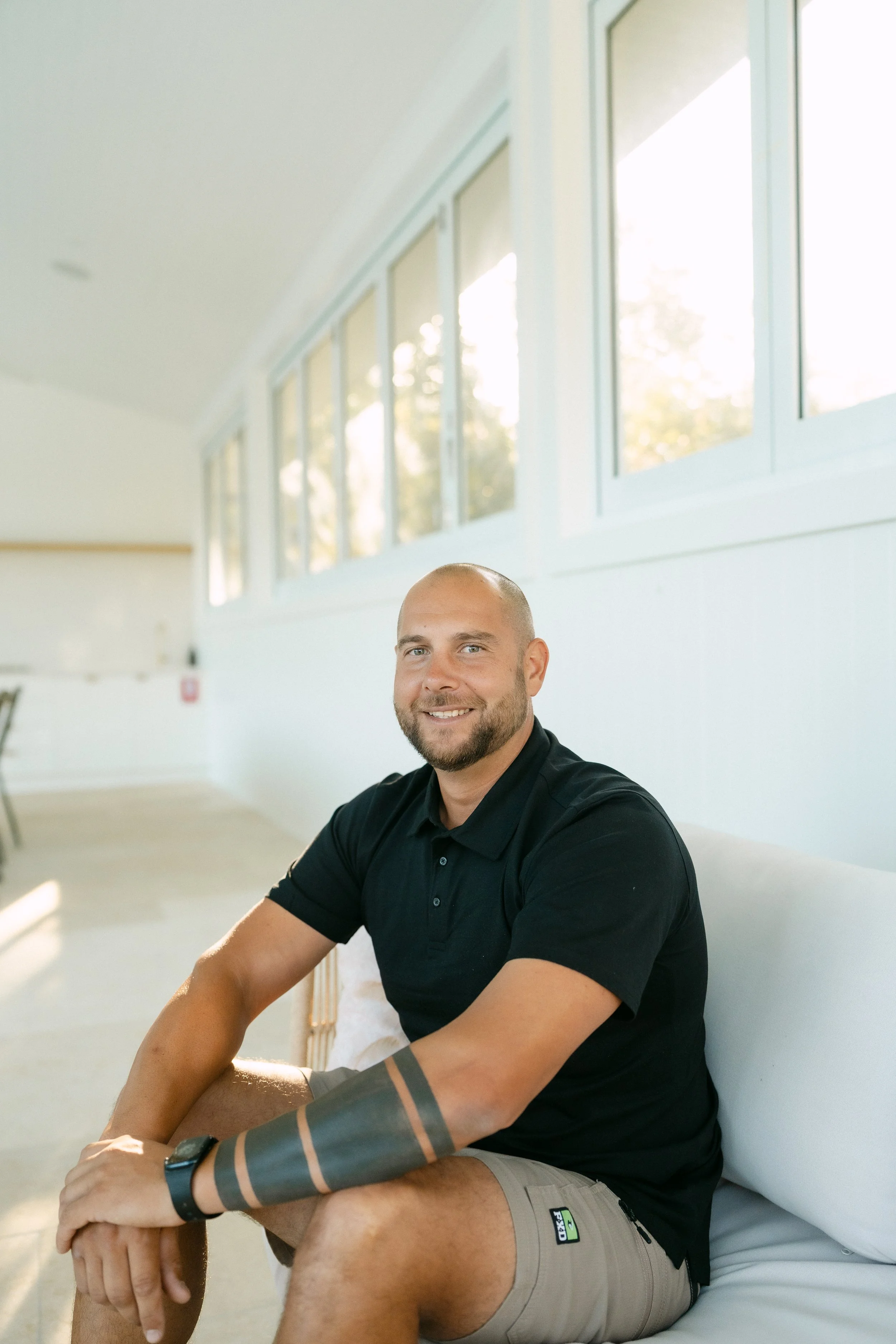 A smiling man with a beard and short hair sitting on a couch in a bright room with large windows, wearing a black polo shirt and khaki shorts, with a tattoo on his left arm.