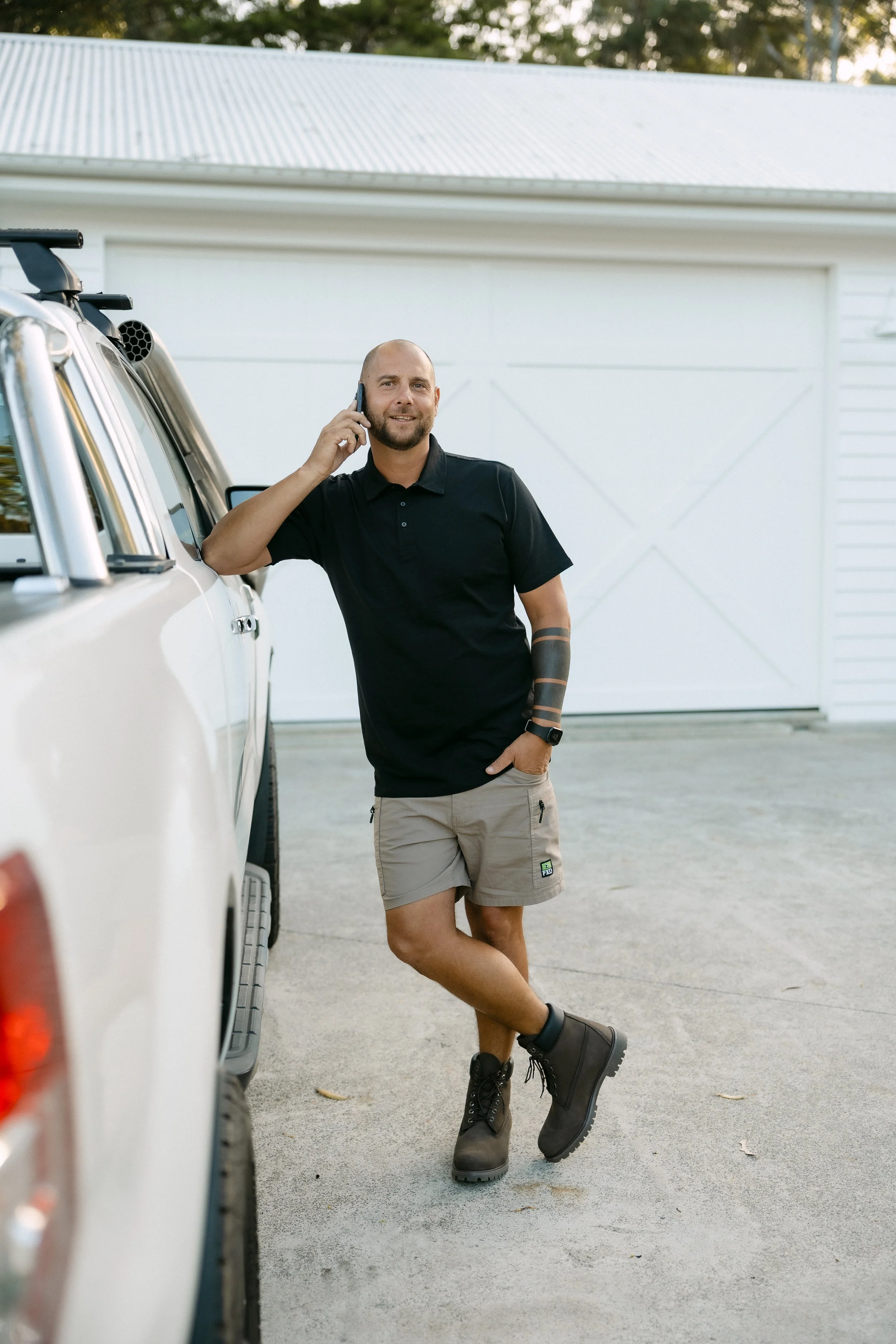 A man with a beard talking on a cell phone while leaning against a white vehicle in a driveway, wearing a black polo shirt, beige shorts, and black boots.