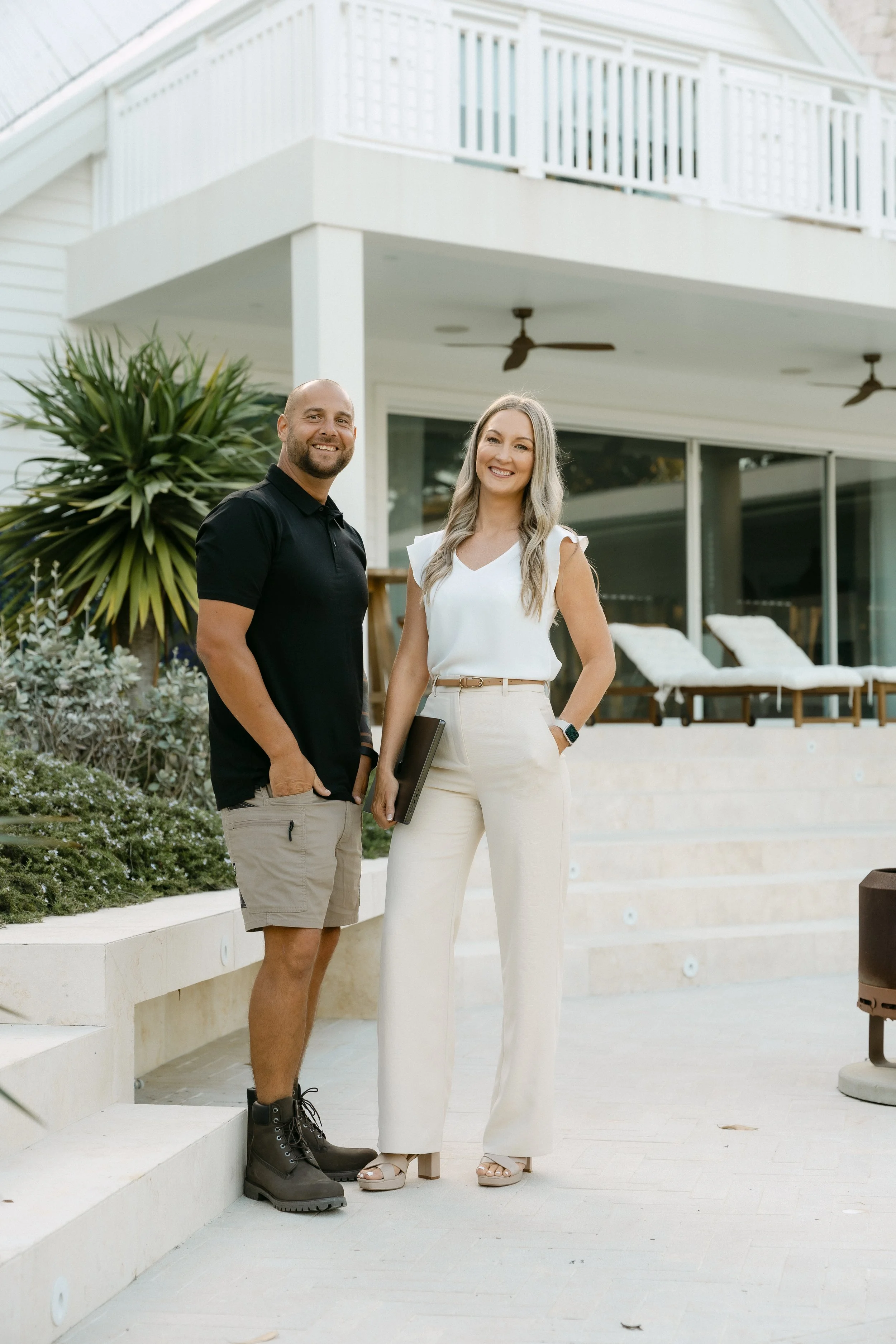 A man and a woman standing outdoors in front of a modern house with a balcony, smiling at the camera, with lush greenery and lounge chairs in the background.