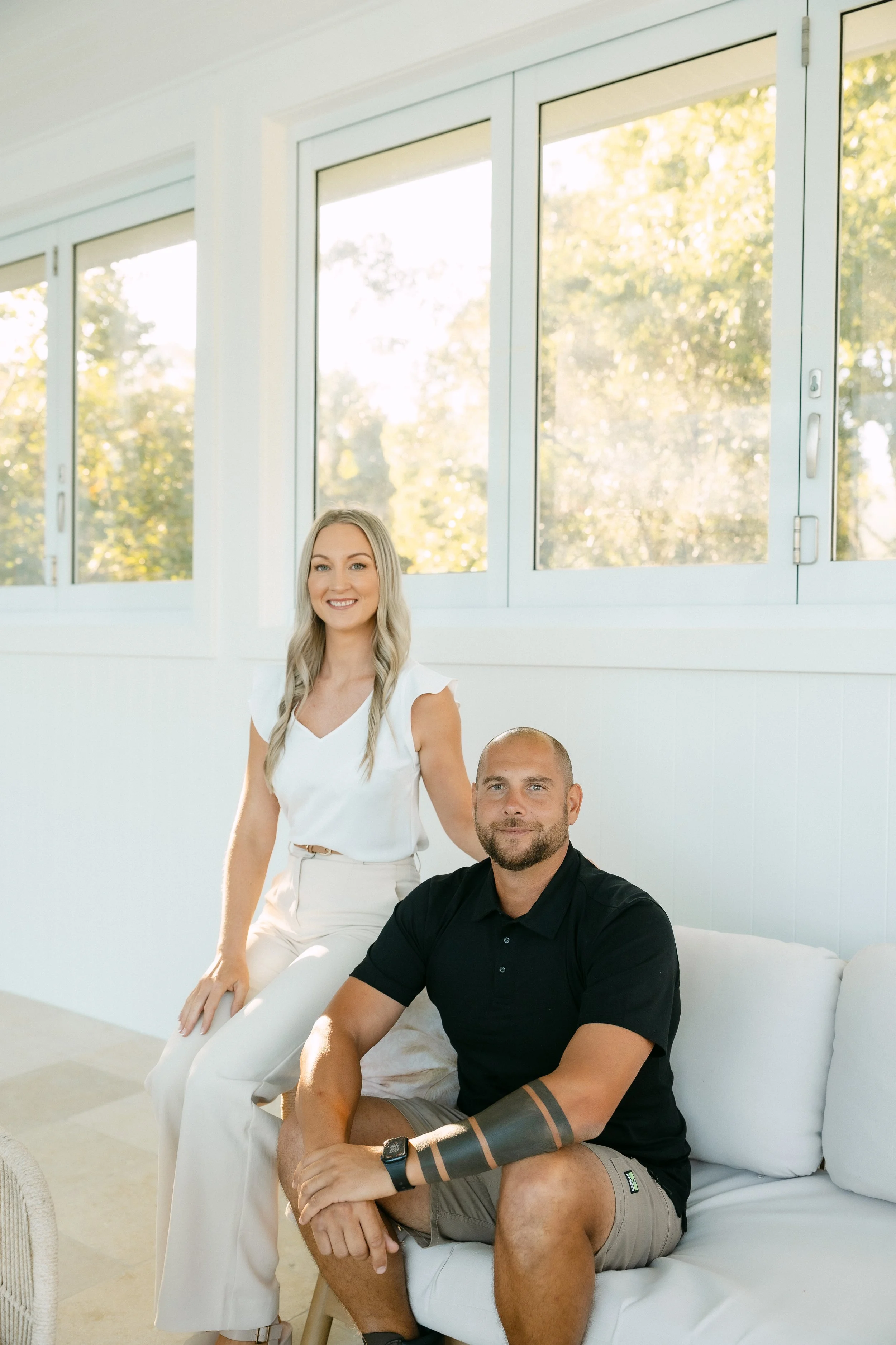 A woman with blonde hair standing next to a seated man with short hair. They are in a bright room with large windows showing trees outside, and they are dressed casually.
