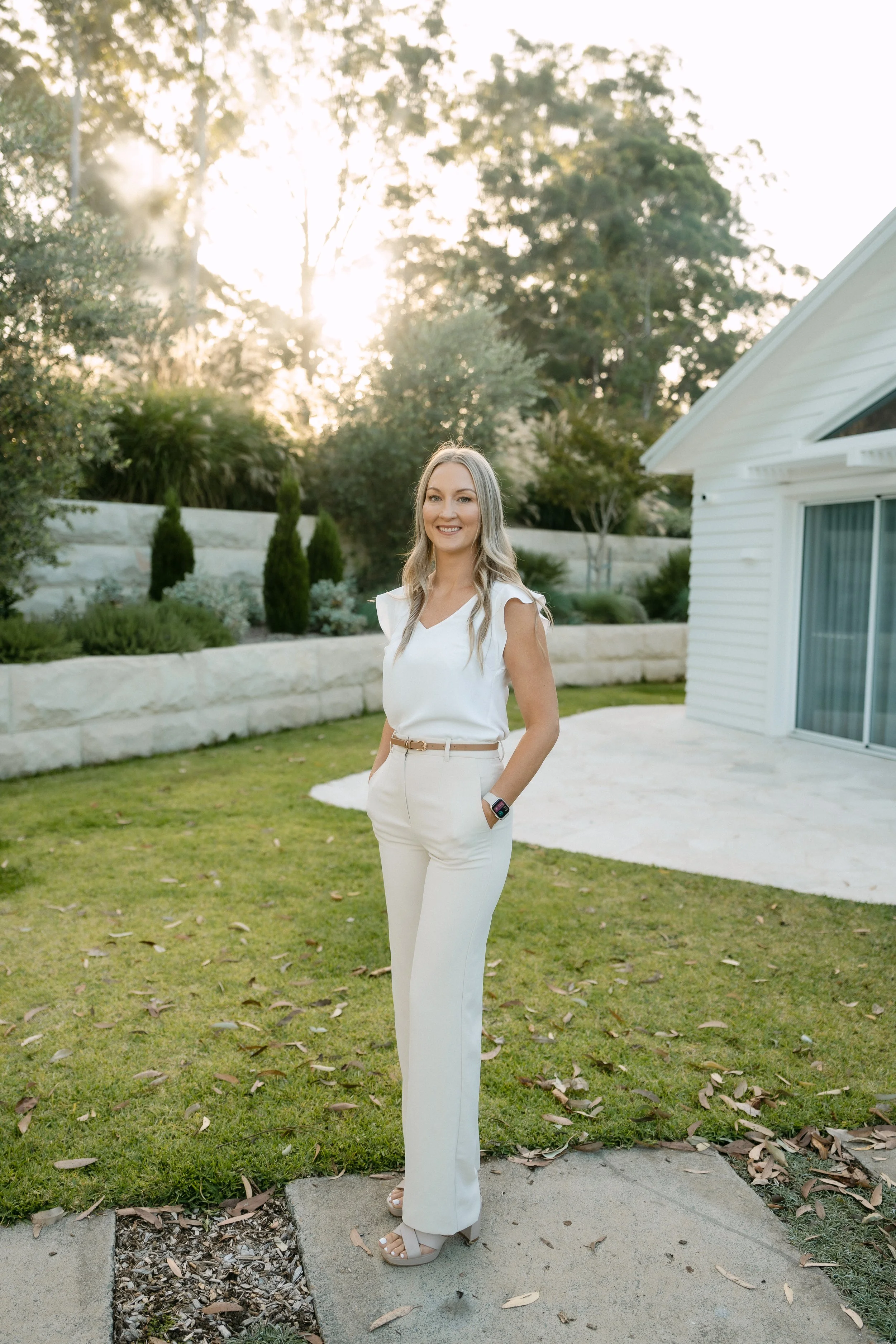 A woman with long blonde hair smiling outdoors in front of a house with a garden, wearing white pants, a white top, and beige high heels.