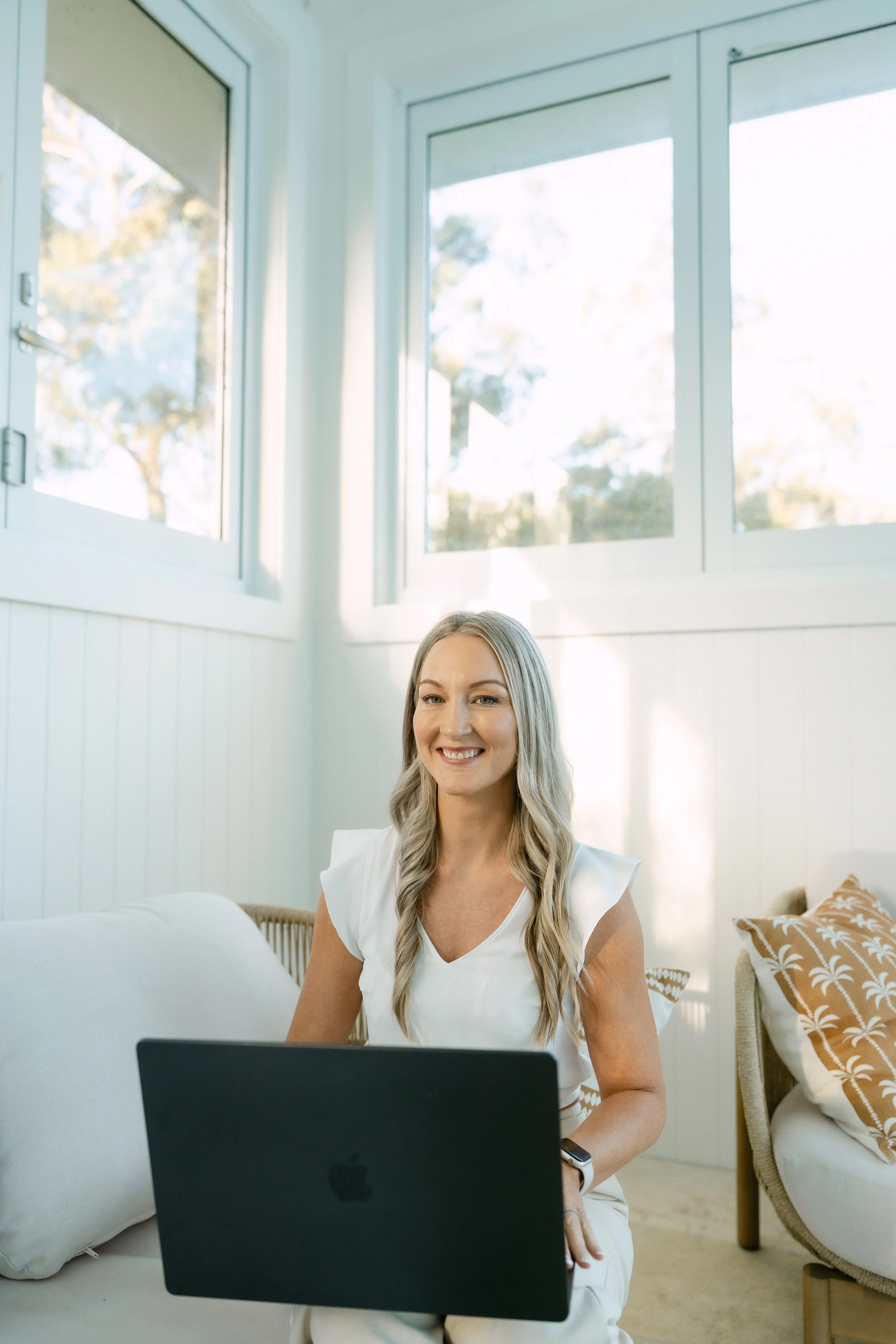 A woman with long blonde hair sitting on a white bed, looking at a laptop, with a bright and cozy room with large windows and patterned pillows behind her.