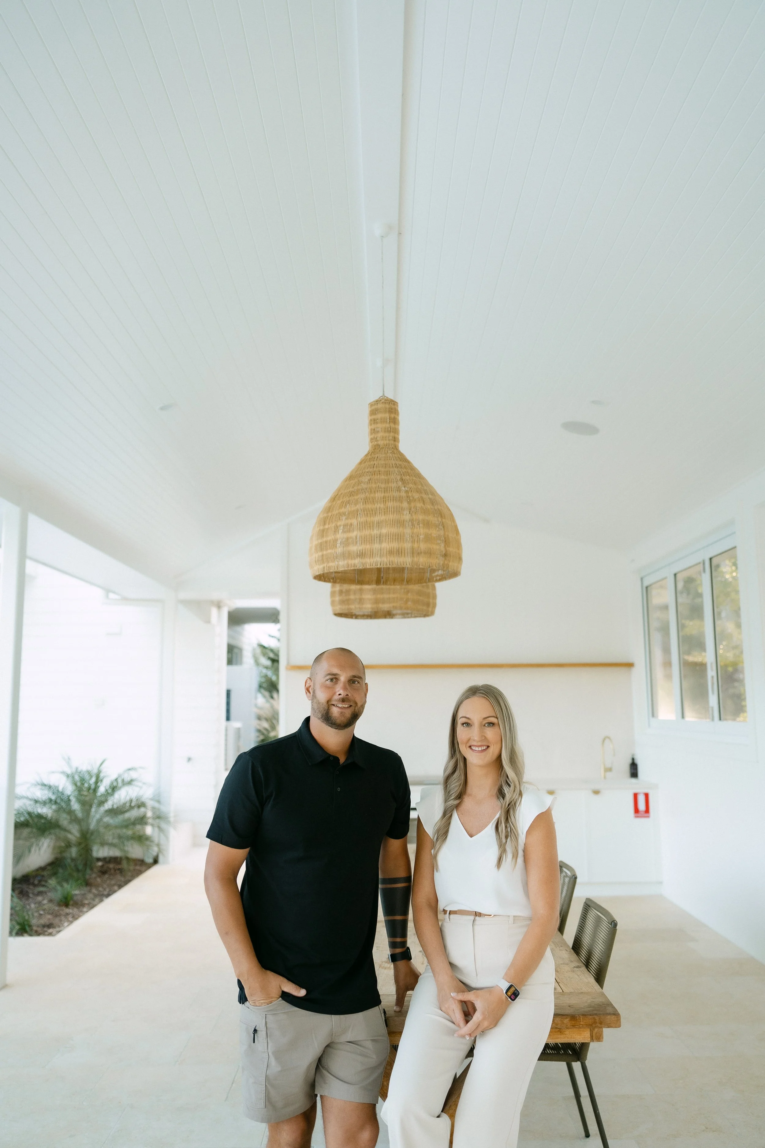 Two people standing in a bright, modern indoor space with large windows, a hanging wicker light fixture, and a wooden table with metal chairs. One man has a beard and is wearing a black polo shirt and light shorts. A woman with long blonde hair is wearing a white top and light-colored pants. They are smiling and looking at the camera.