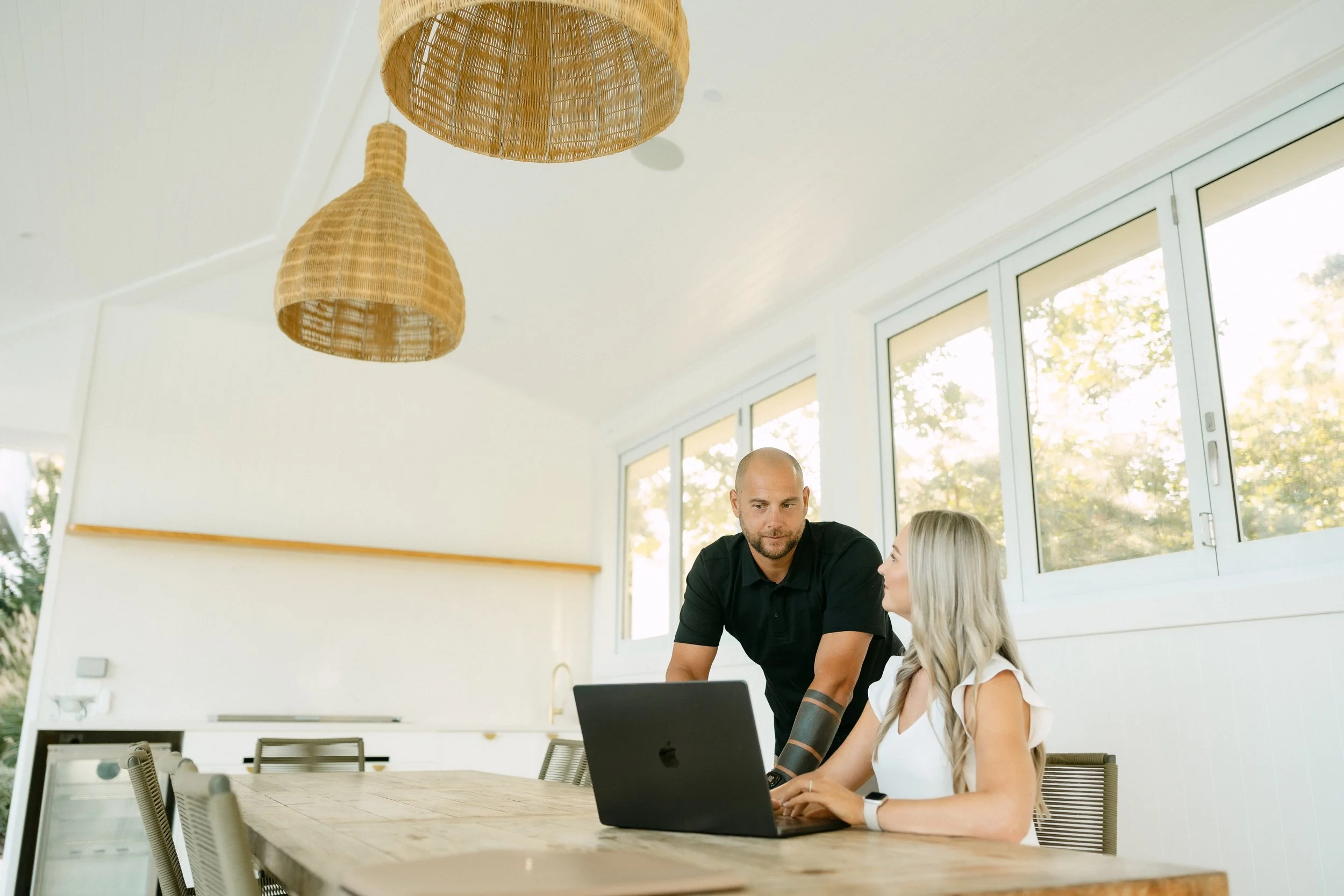 Two people, a man and a woman, work together on a laptop in a bright, modern office with large windows and hanging woven light fixtures.