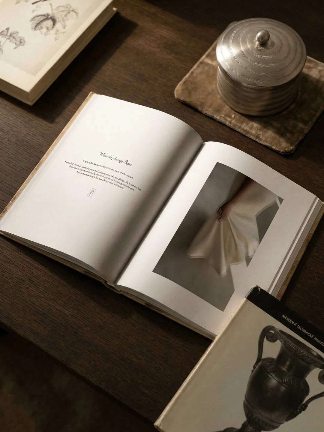 An open book displaying text and a photograph of a white wedding dress, a closed book about vases, and a decorative metal container on a wooden table.