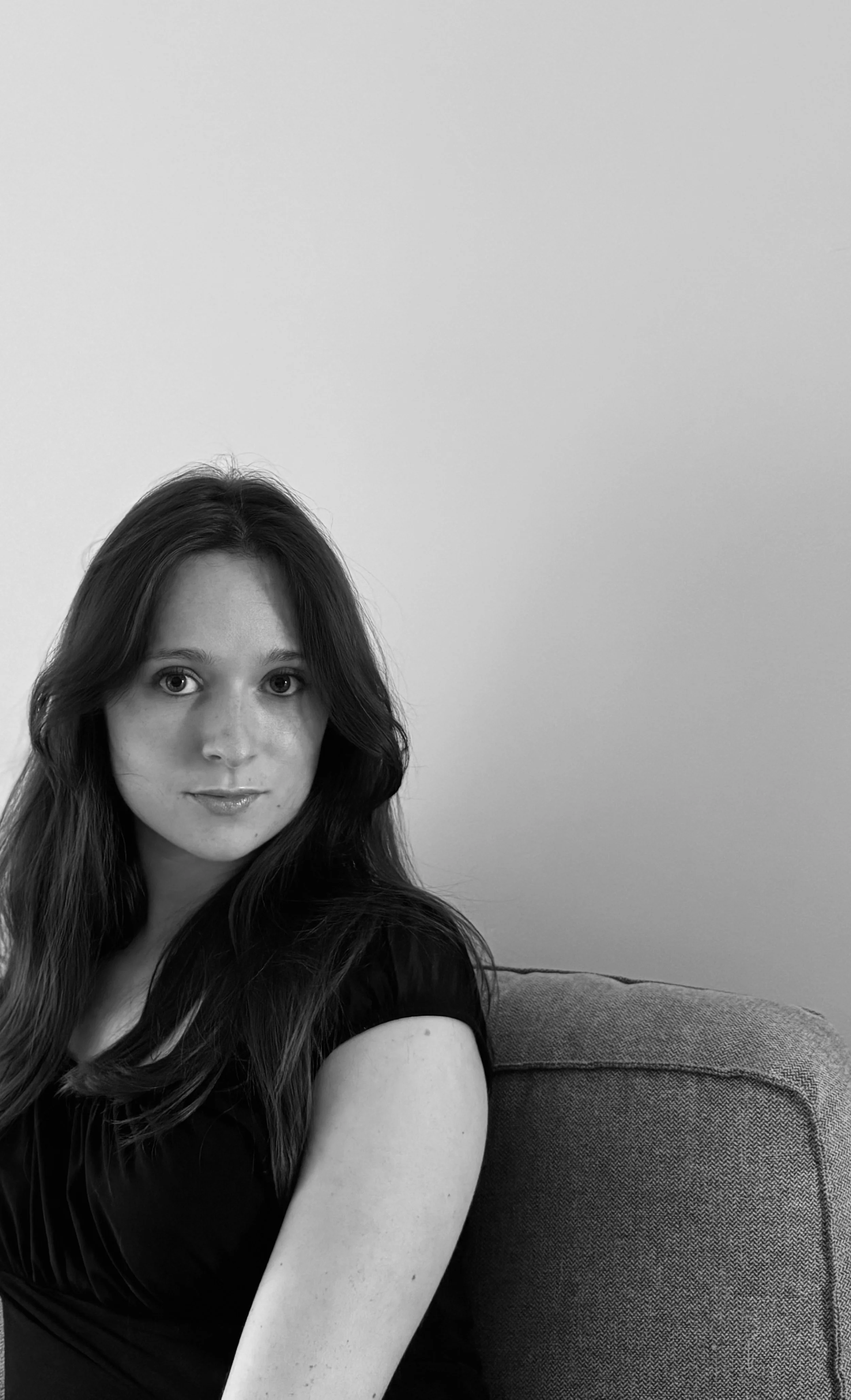 Une femme avec de longs cheveux foncés, portant un haut noir, assise sur un canapé, regardant vers l'objectif, en noir et blanc.