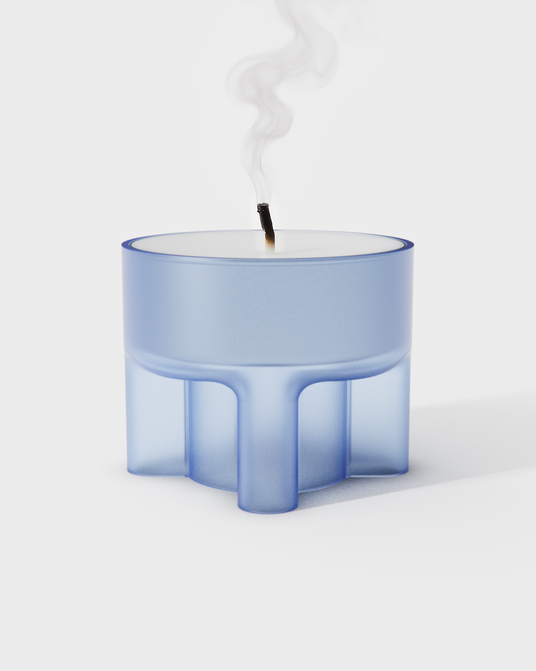 A lit candle with smoke rising from the wick, housed in a modern, frosted blue glass holder with a geometric design, on a white surface with a plain white background.