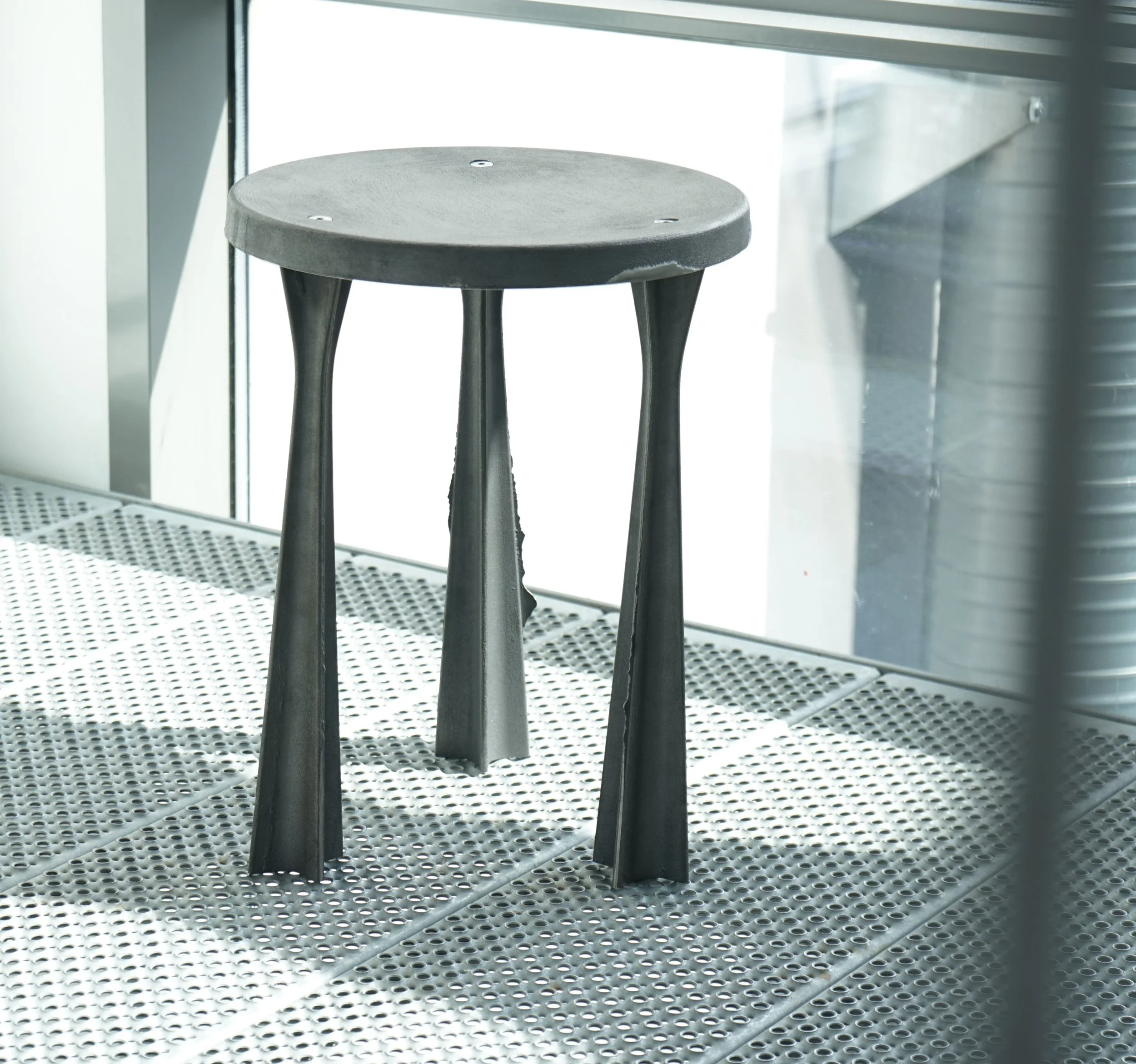 A small, round, black stool with three legs is positioned on a perforated metal surface near a glass window or door, with sunlight illuminating part of the scene.