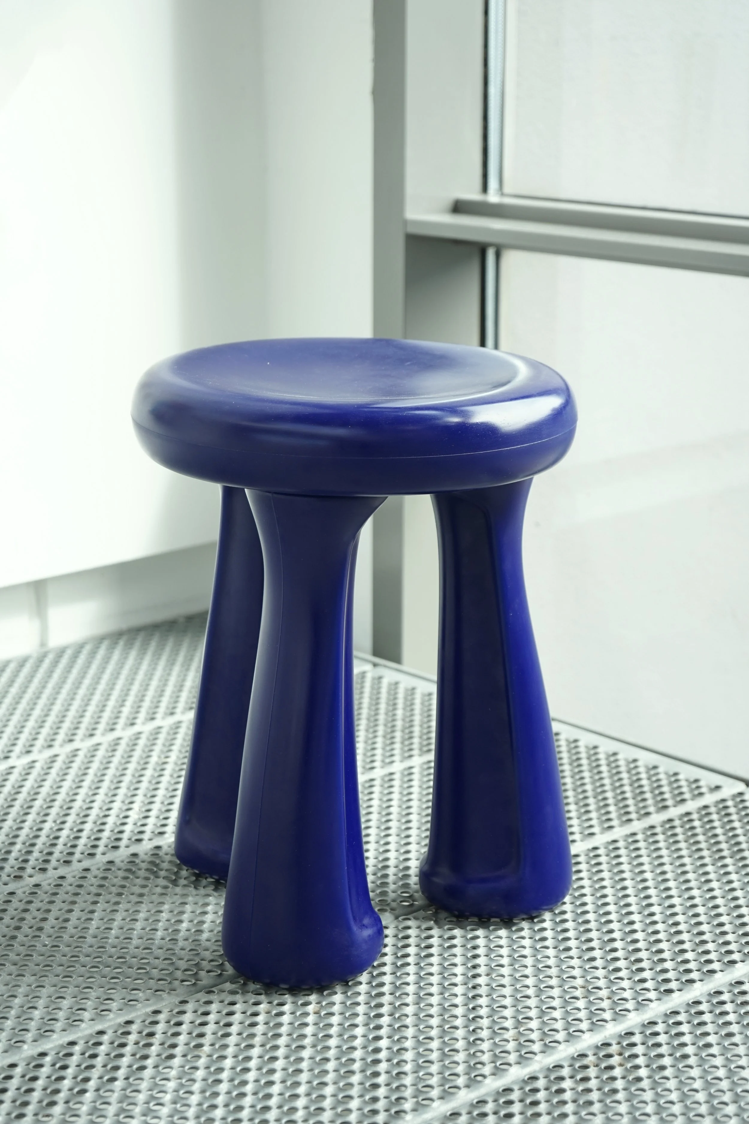 Close-up of a dark blue plastic stool with three legs, positioned on a perforated metal surface near a window with a white wall in the background.