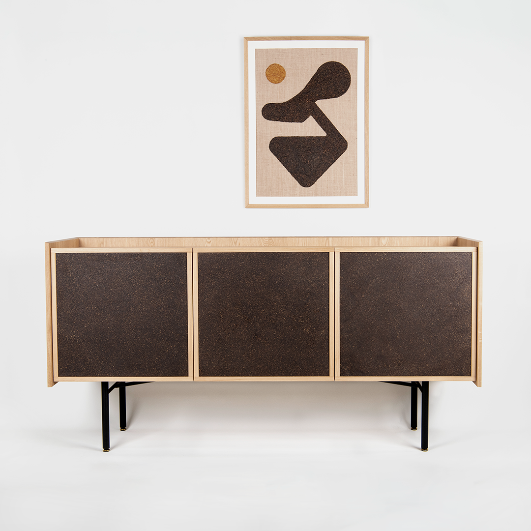 A wooden sideboard with dark doors standing against a white wall, with abstract artwork hanging above it that features a large dark shape, a small brown circle, and a pink background.