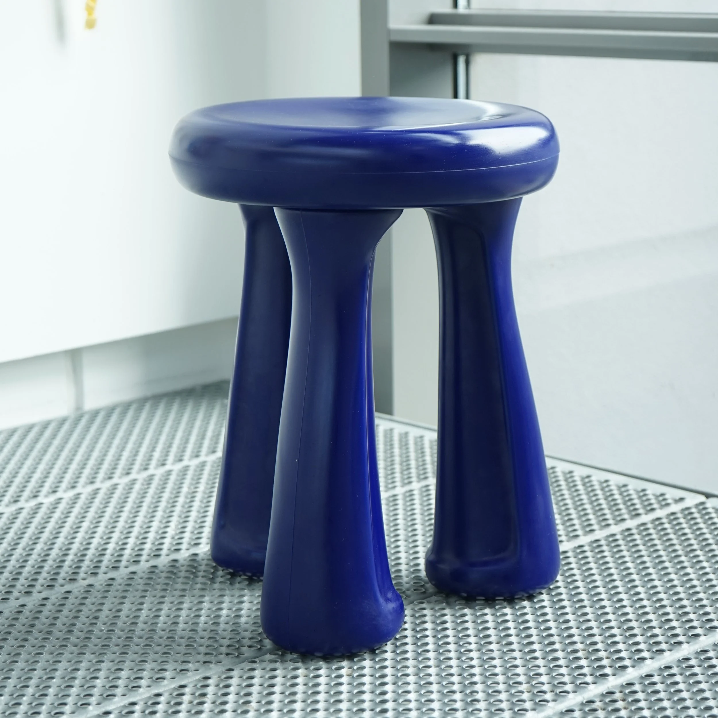 Close-up of a blue stool with three legs on a perforated gray metal floor near a white wall and furniture.