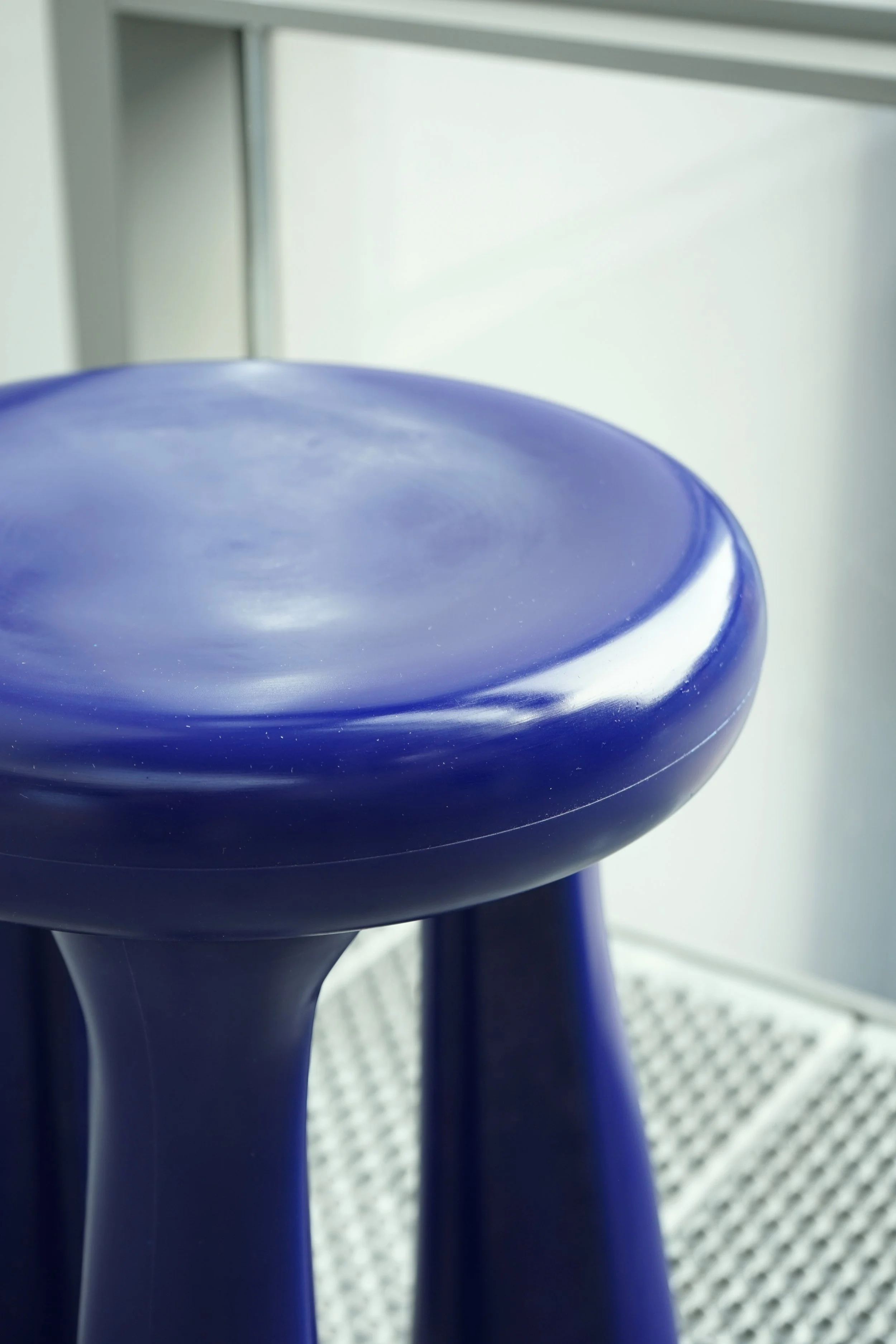 Close-up of a blue stool with a rounded seat and three curved legs, positioned on a patterned rug near a white cabinet.