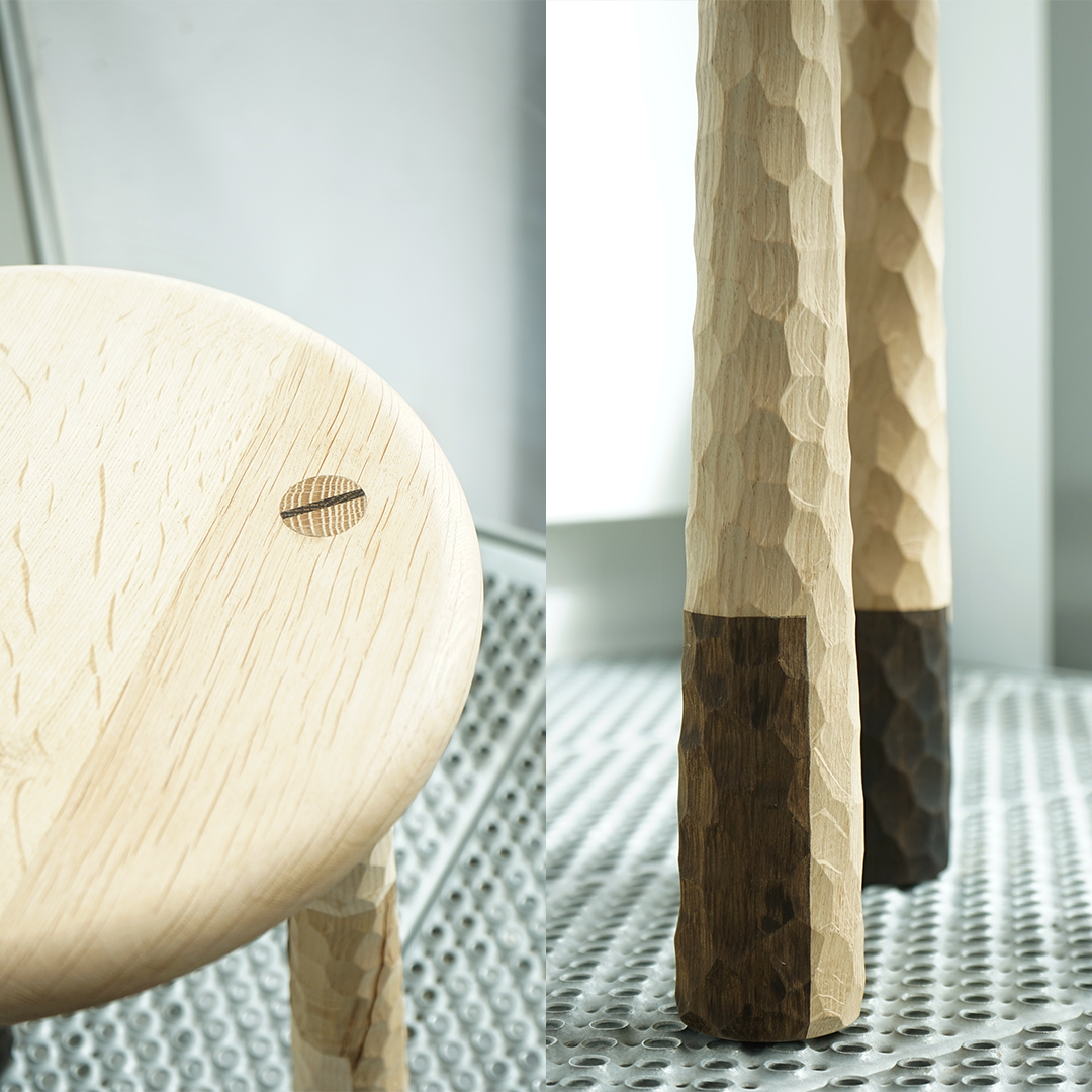 Close-up of a light-colored wooden table with a screw on the surface and a carved, part of a wooden leg, showing a textured, honeycomb-like pattern. The table stands on a perforated metal surface near a window.