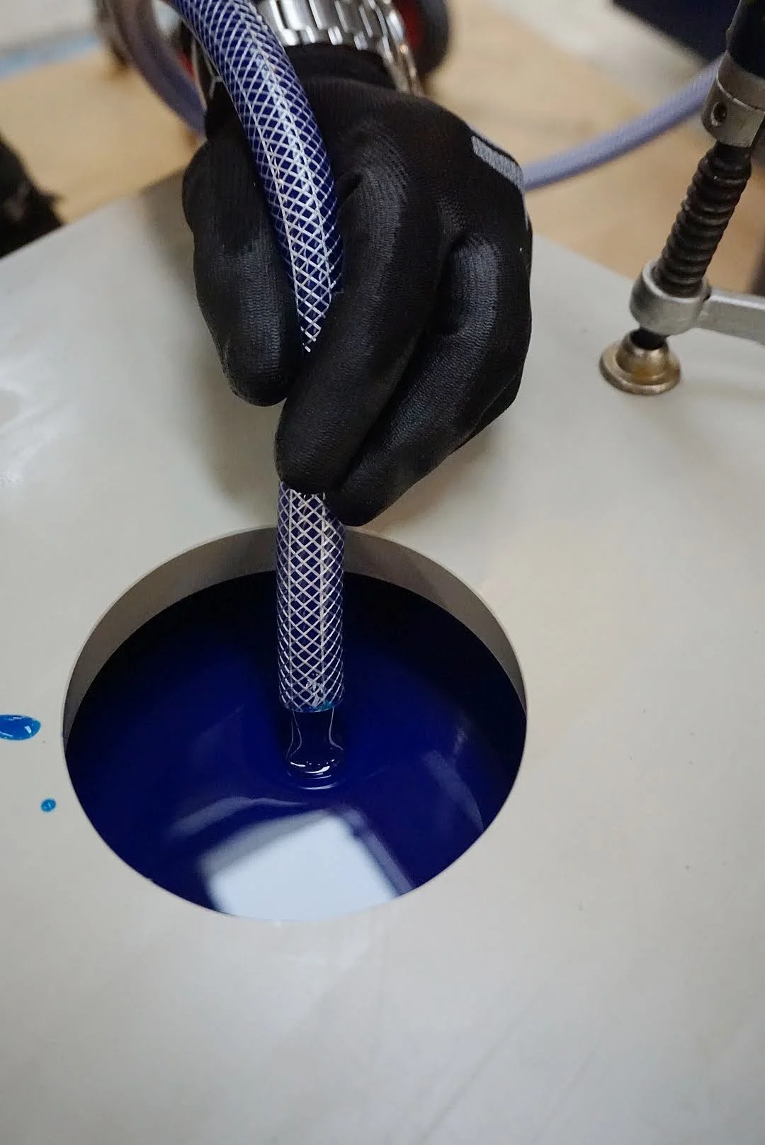 A person wearing a black glove is inserting a braided blue and white cable into a hole on a device. The device has a black circular opening with a blue liquid inside. Part of a metallic spring and a gray hose are visible on the right side.