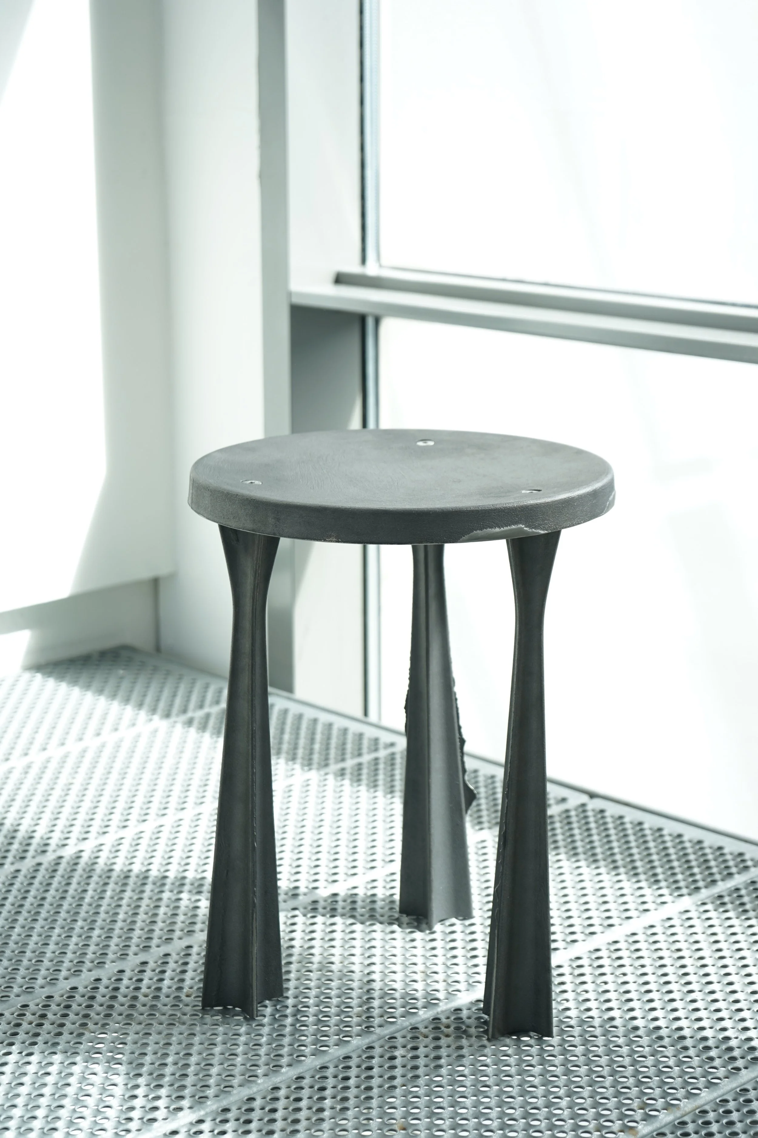 A black stool with three curved legs, placed on a perforated white metal floor near a window with white curtains.