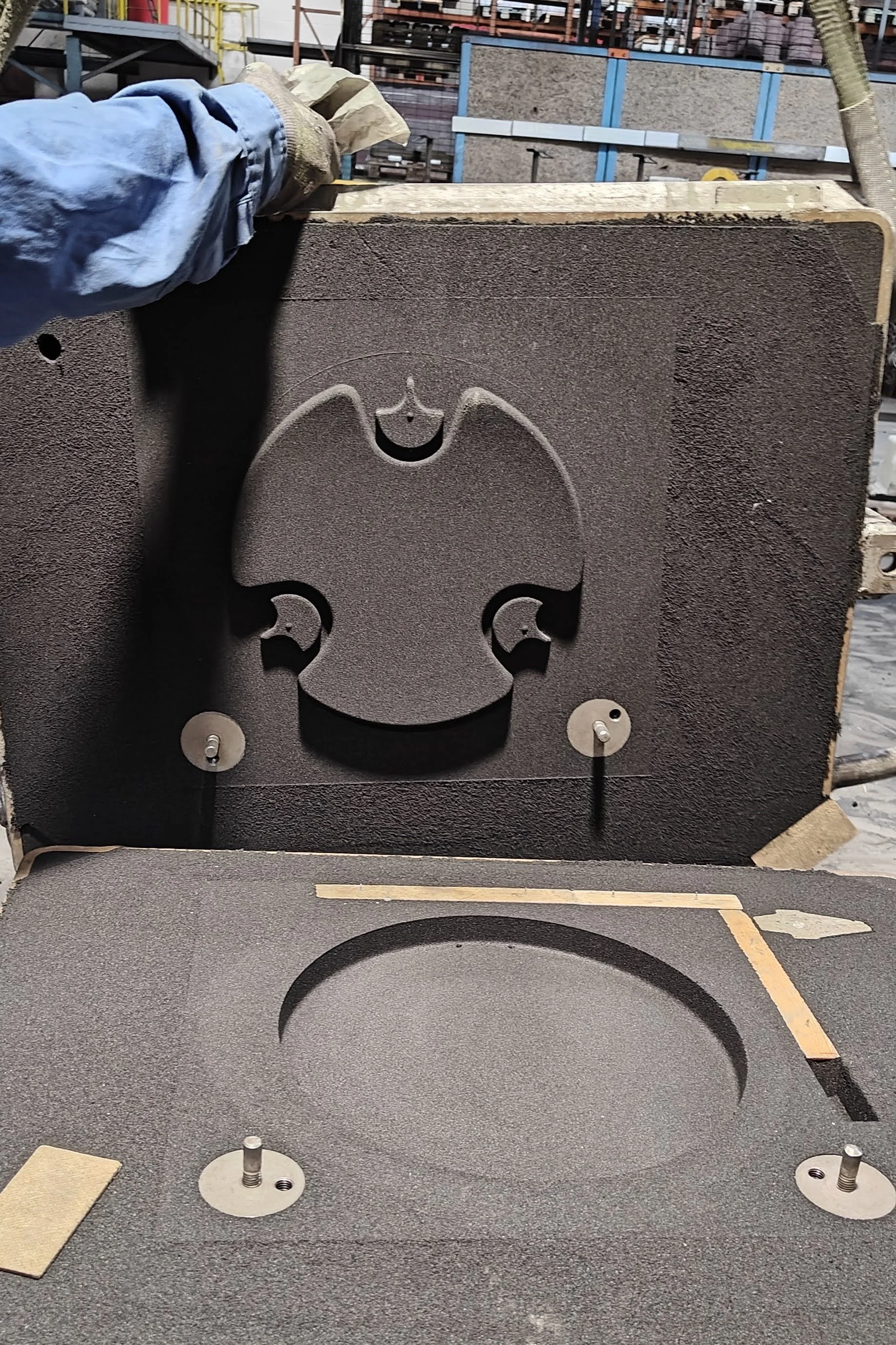 A workshop showing a large black piece of machinery part with a skull-shaped cutout. The piece is mounted on a table with scattered small equipment and tools nearby.