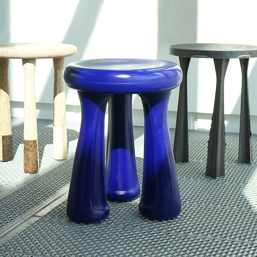 Three stools with different designs and colors on a metal grid floor, sunlight streaming through a window in the background.