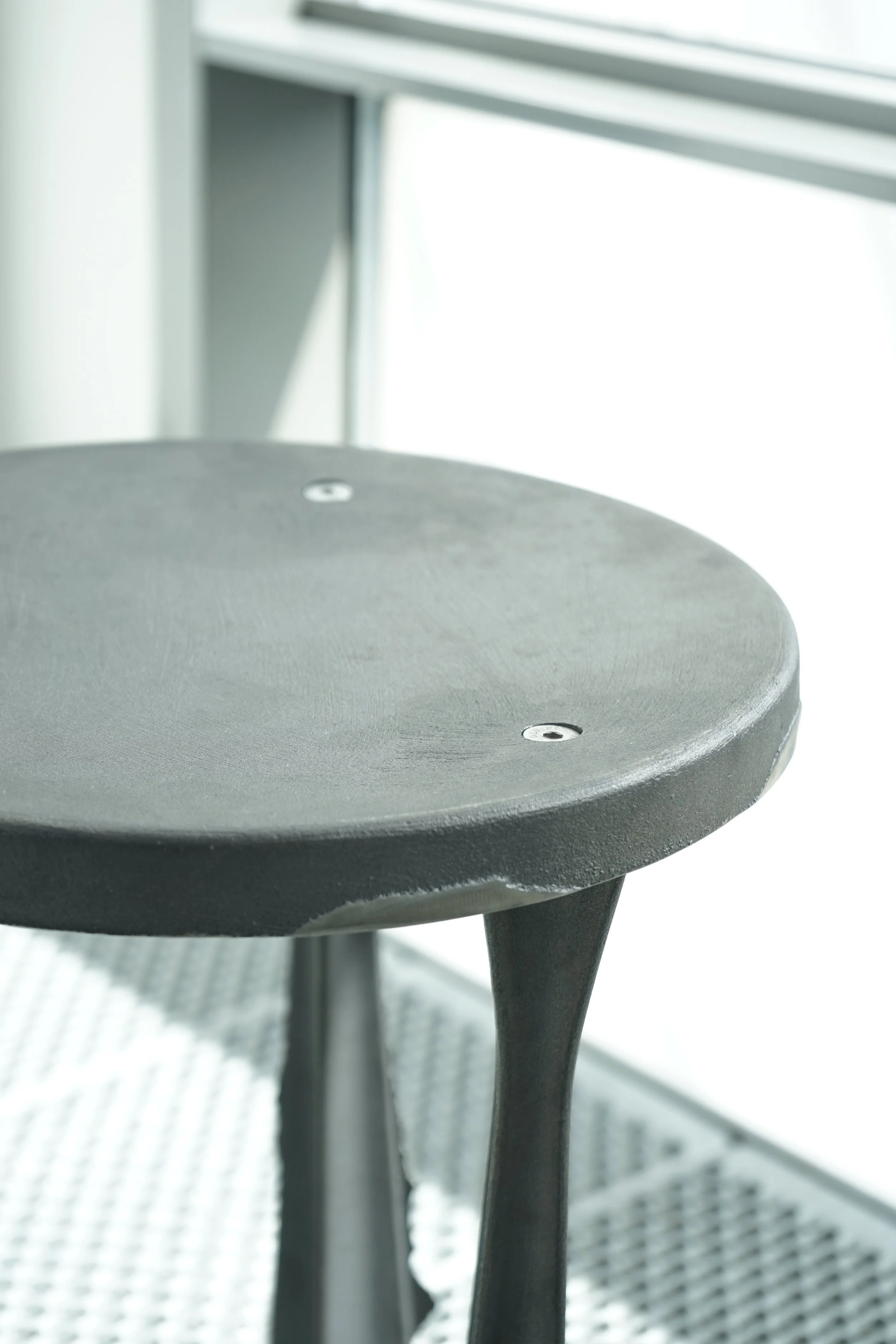 Close-up of a black stool with a round seat and wooden legs, adjacent to a window with blinds.