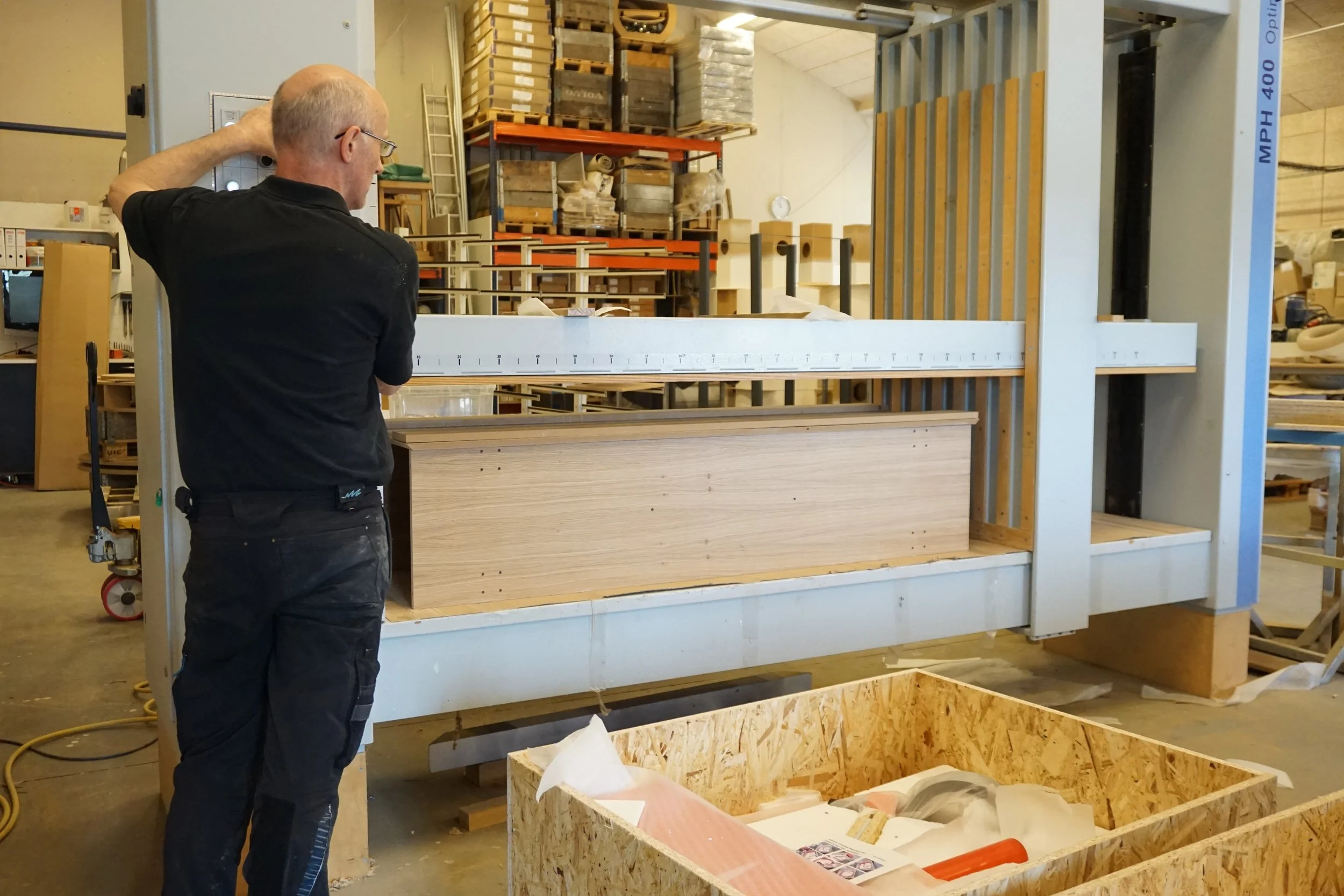 A man with glasses and a black shirt is inspecting a large wooden frame or structure in a workshop or storage area, with shelves full of boxes and materials in the background.