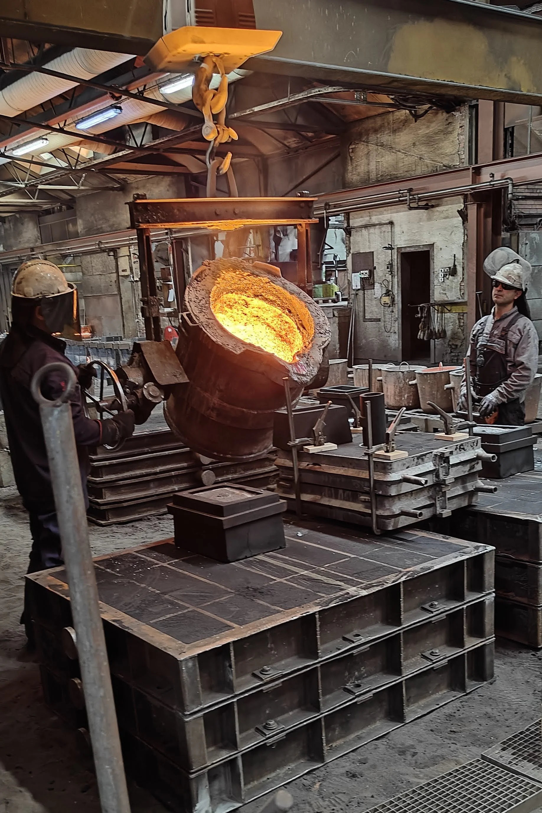 A steel mold being filled with molten metal in a foundry, with workers wearing protective gear including helmets and face shields, and a furnace in the background.