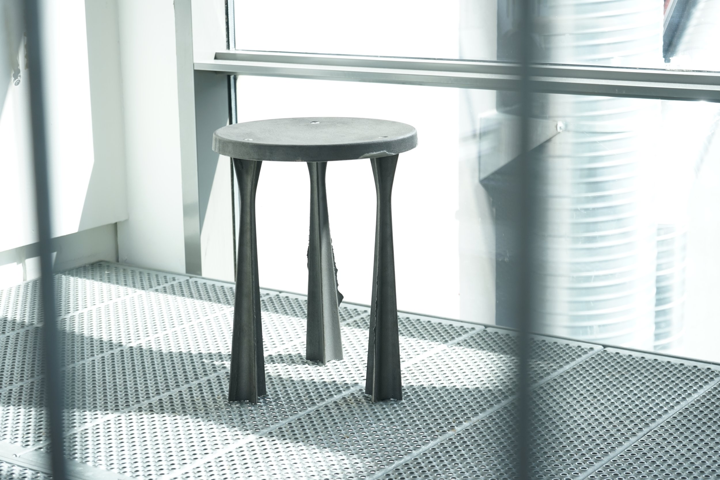 A black wooden stool with three legs inside a bright room with large windows and metal vent pipes, seen through a window screen.