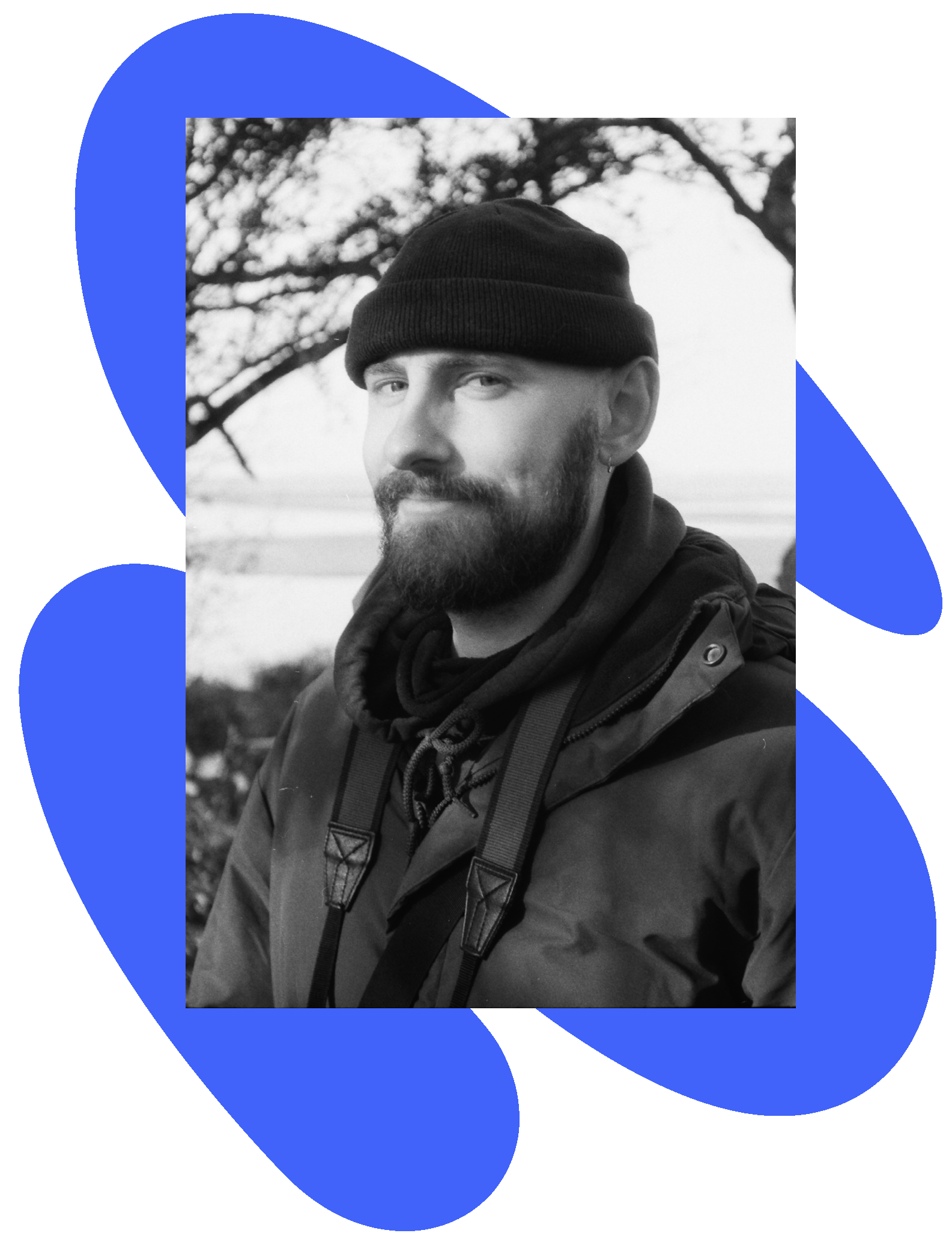 Black and white photo of a man with a beard, wearing a beanie and a jacket, outdoors with trees in the background.