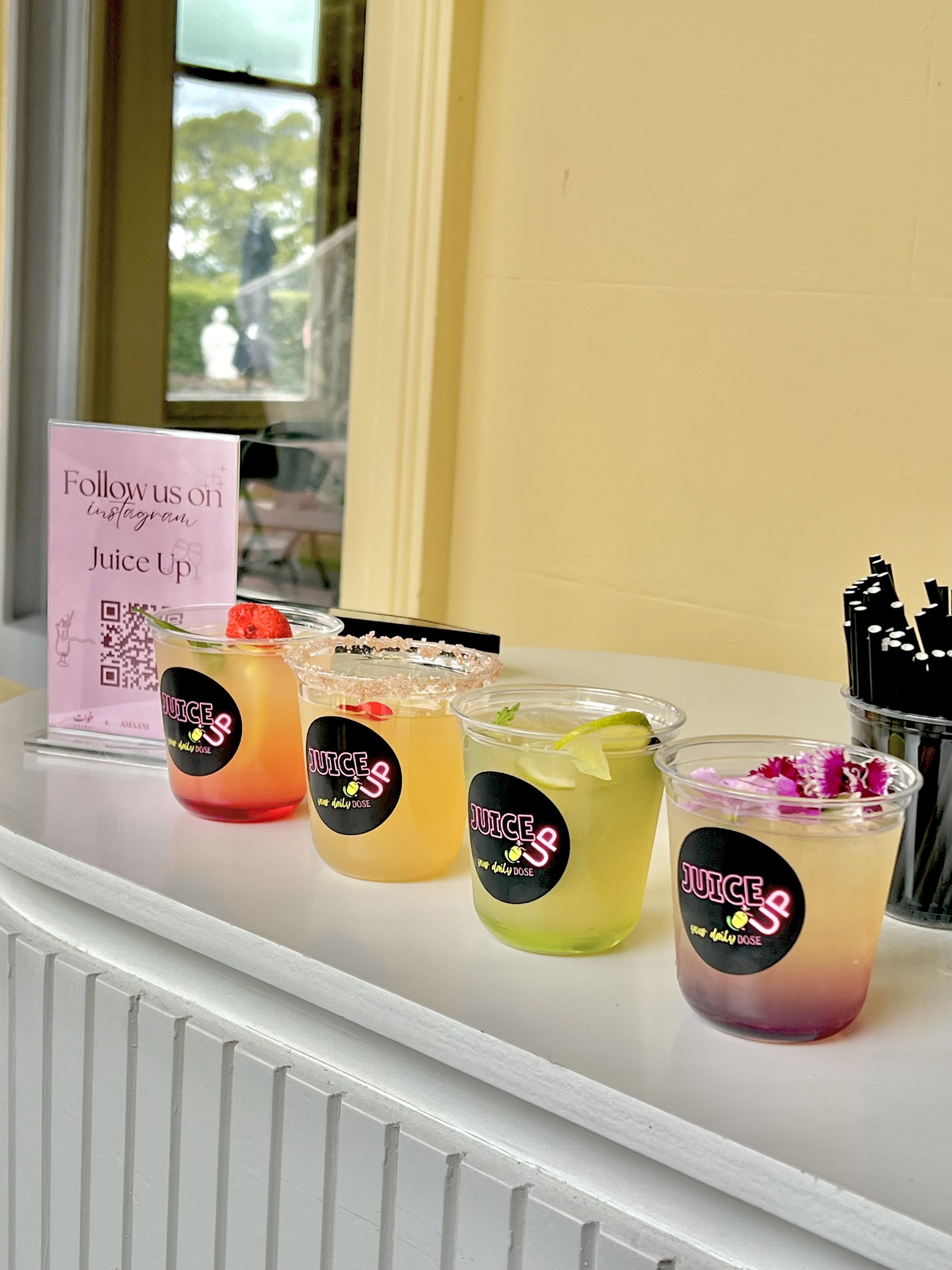 Colorful beverages in plastic cups with black stickers labeled 'Juice Up' on a white countertop. A pink sign promotes following on Instagram for Juice Up. The drinks have various garnishes like lime slices, a strawberry, and flowers. In the background, a window shows greenery and outdoor scenery.