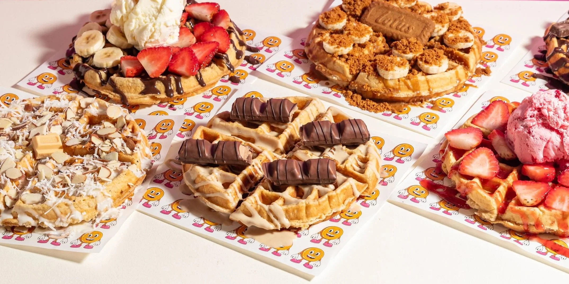 Various waffles topped with strawberries, bananas, chocolate, whipped cream, and other toppings arranged on colorful paper with a smiley face design.