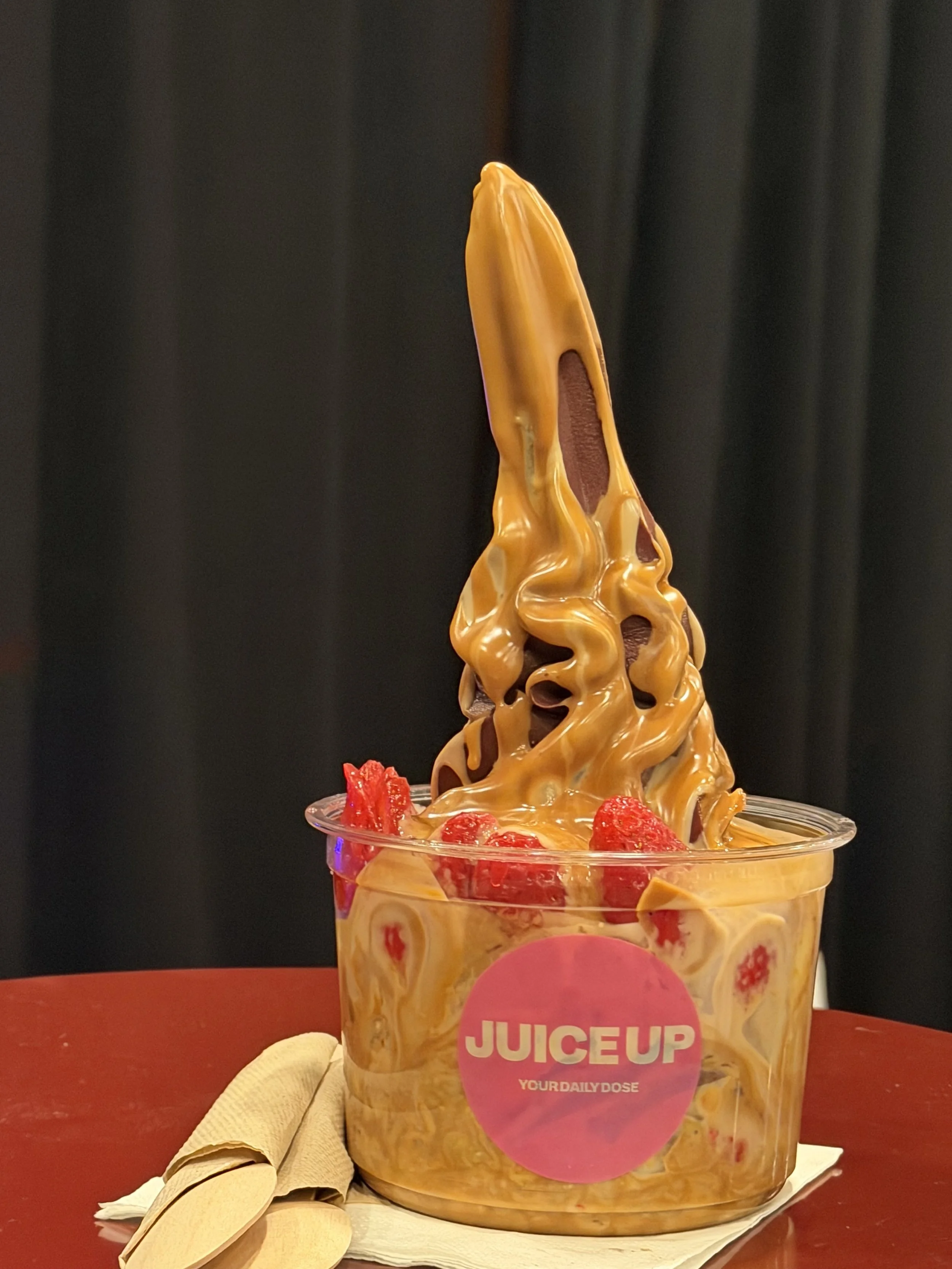 A dessert cup filled with caramel swirl soft serve ice cream, topped with strawberries, on a red table. A beige napkin and a yellow straw wrapper are placed beside the cup.