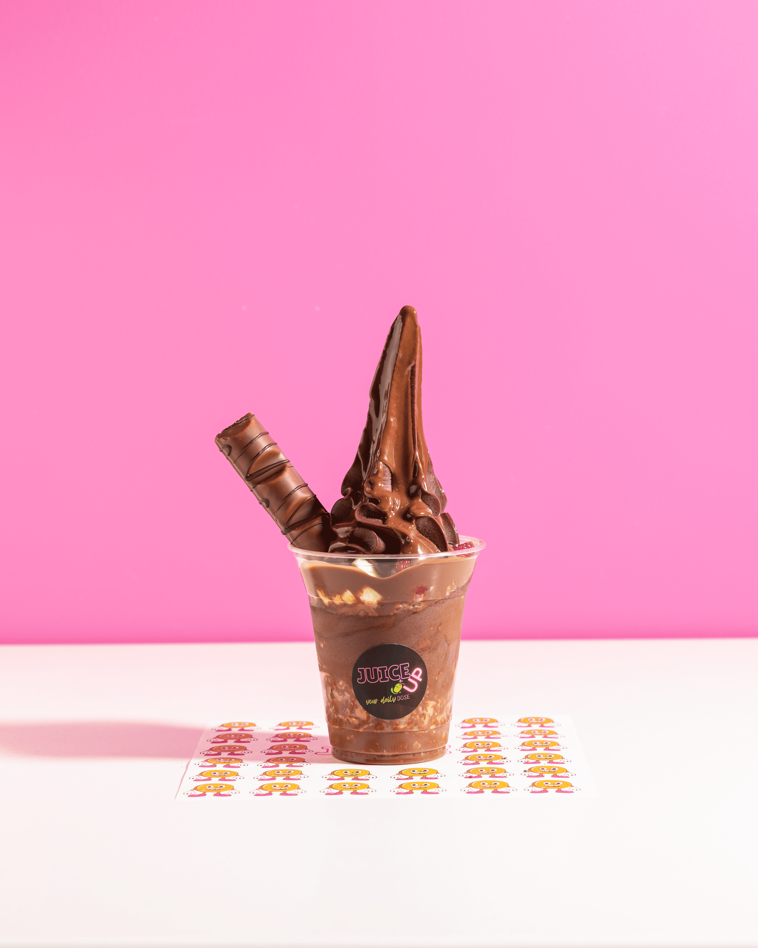 A chocolate soft serve ice cream cone with two chocolate wafer sticks, served in a clear plastic cup with a black sticker that reads 'Juice Up your daily dose', placed on a white surface with colorful stickers, against a pink background.