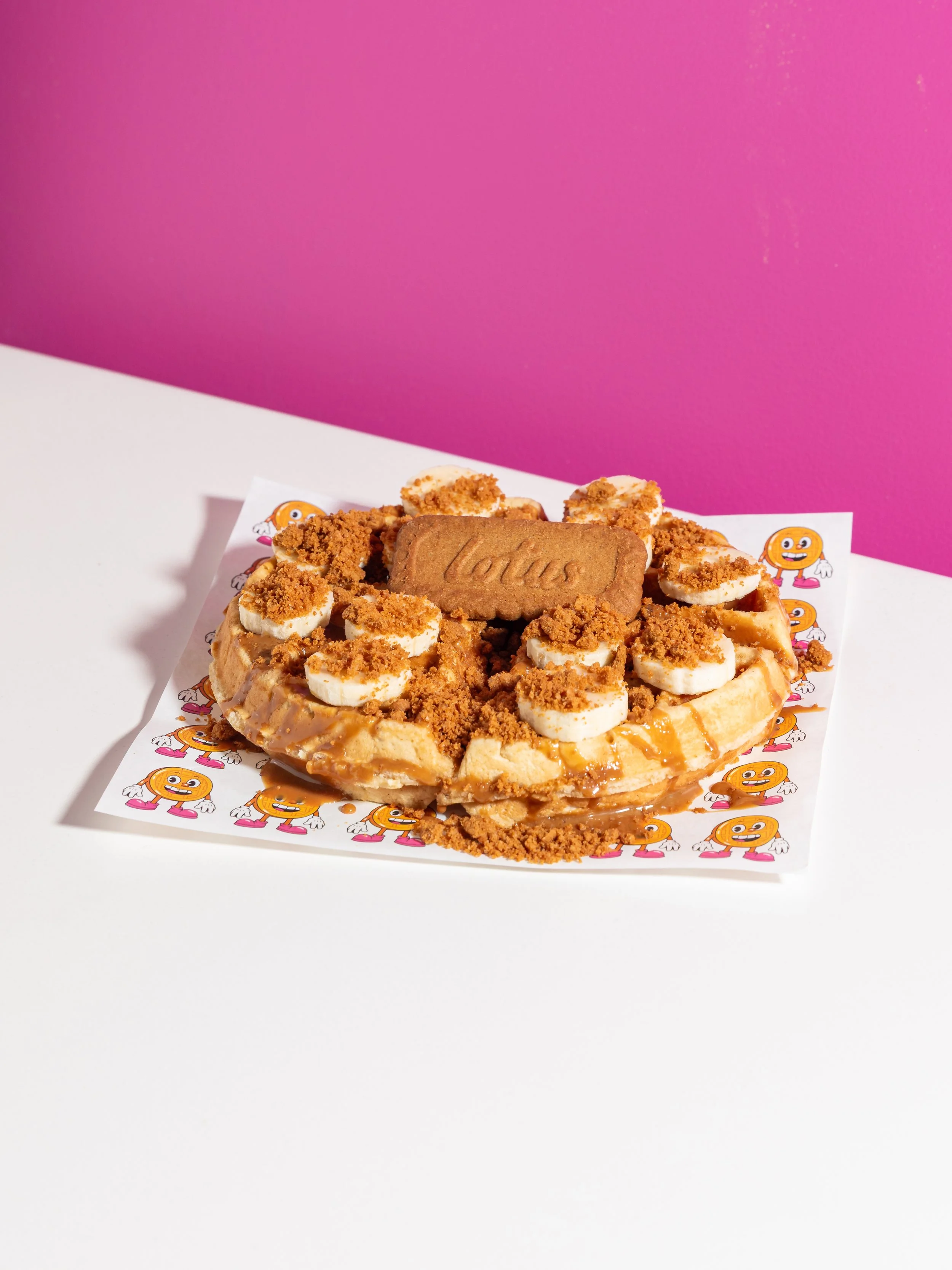 A dessert cake topped with banana slices, caramel sauce, and cookie crumbs, with a Lotus biscuit on top, placed on paper printed with orange cartoon characters, on a white surface with a pink background.