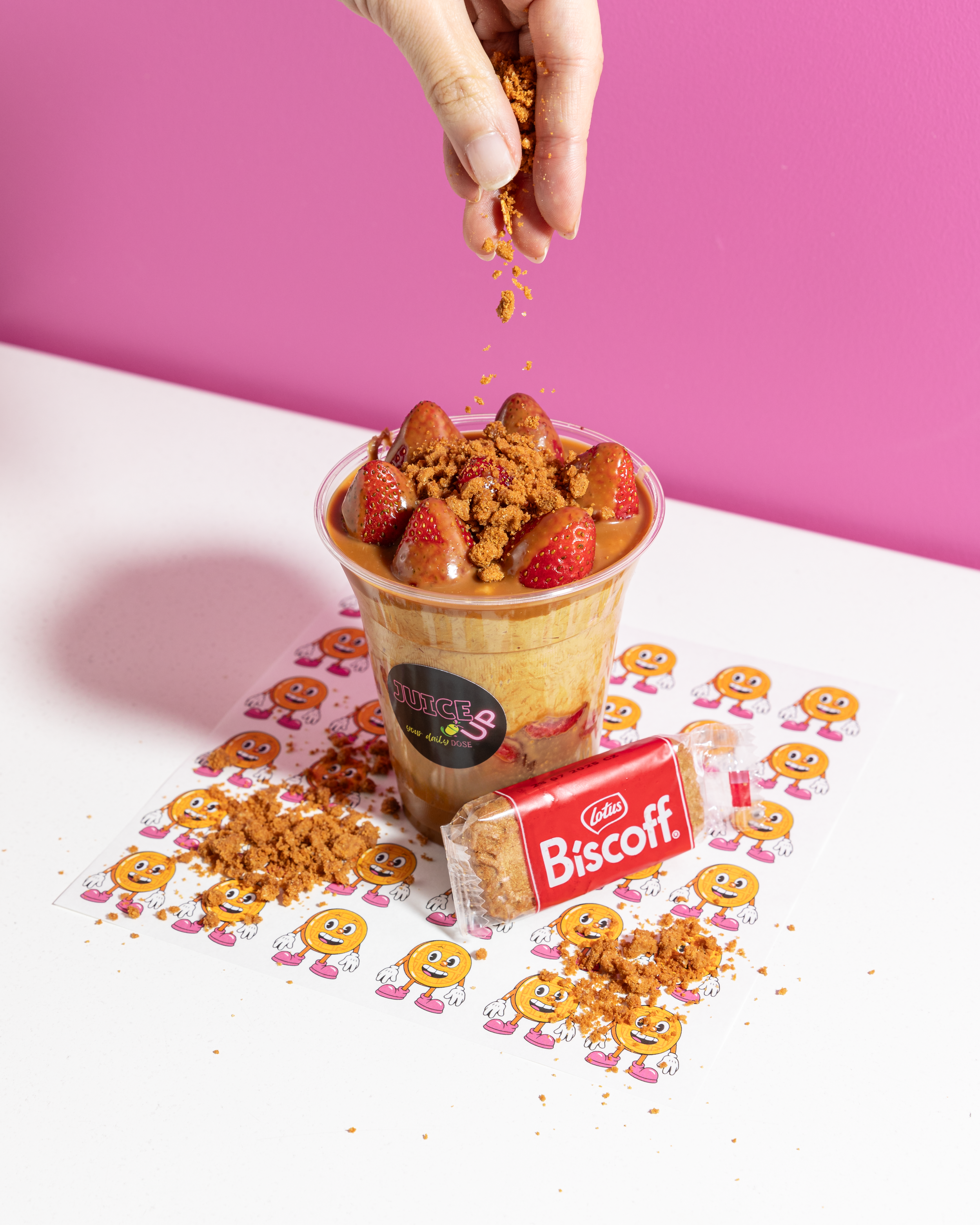 A hand is sprinkling brown crumble over strawberries and a drink in a clear cup on a table, with a pink background and cartoon character print paper underneath.