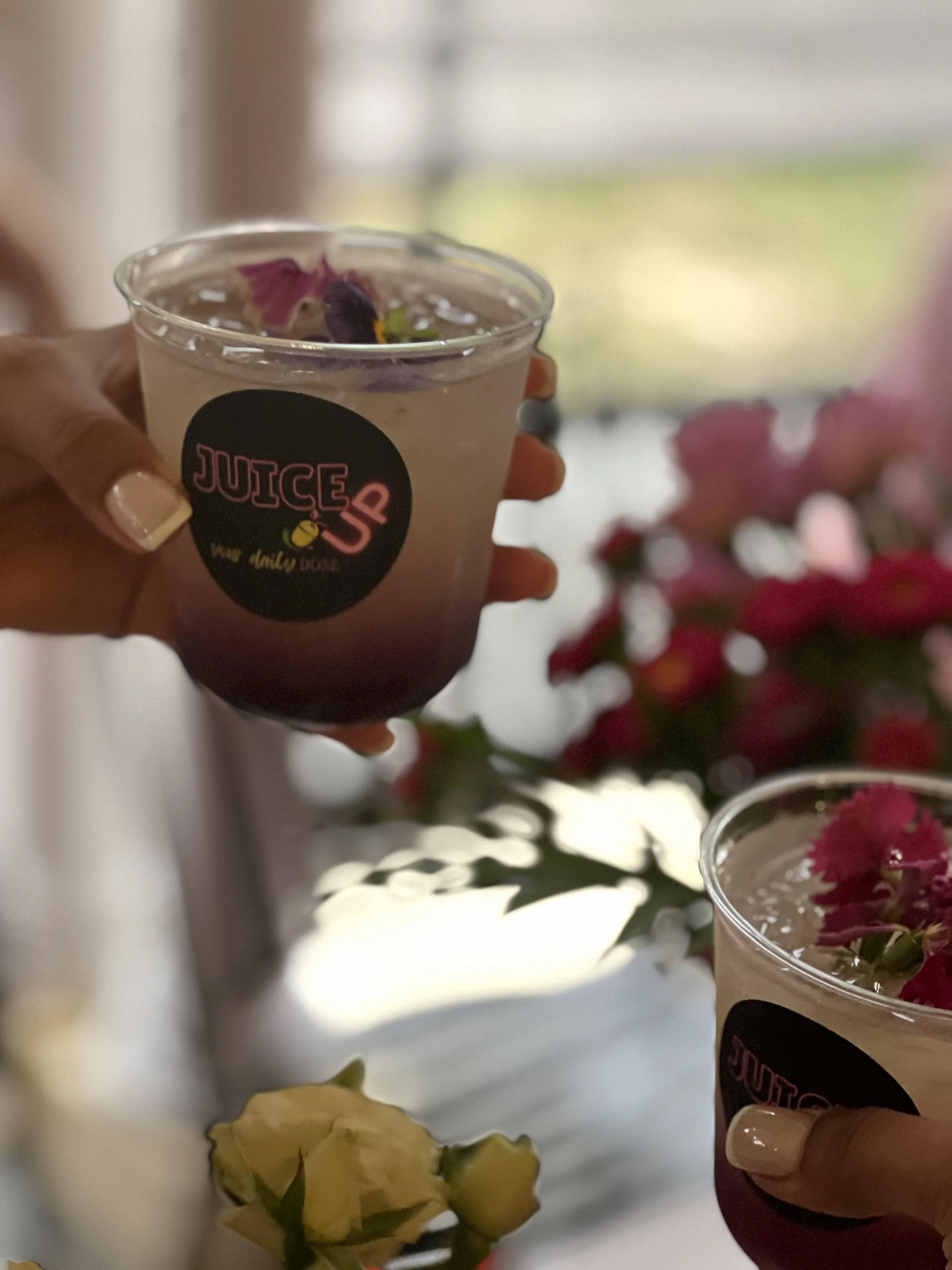 A hand holding a clear plastic cup with a pink and black label reading 'JUICE UP your daily dose.' The cup contains a beverage topped with purple and green edible flowers. In the background, there are pink flowers and yellow flowers on a table.