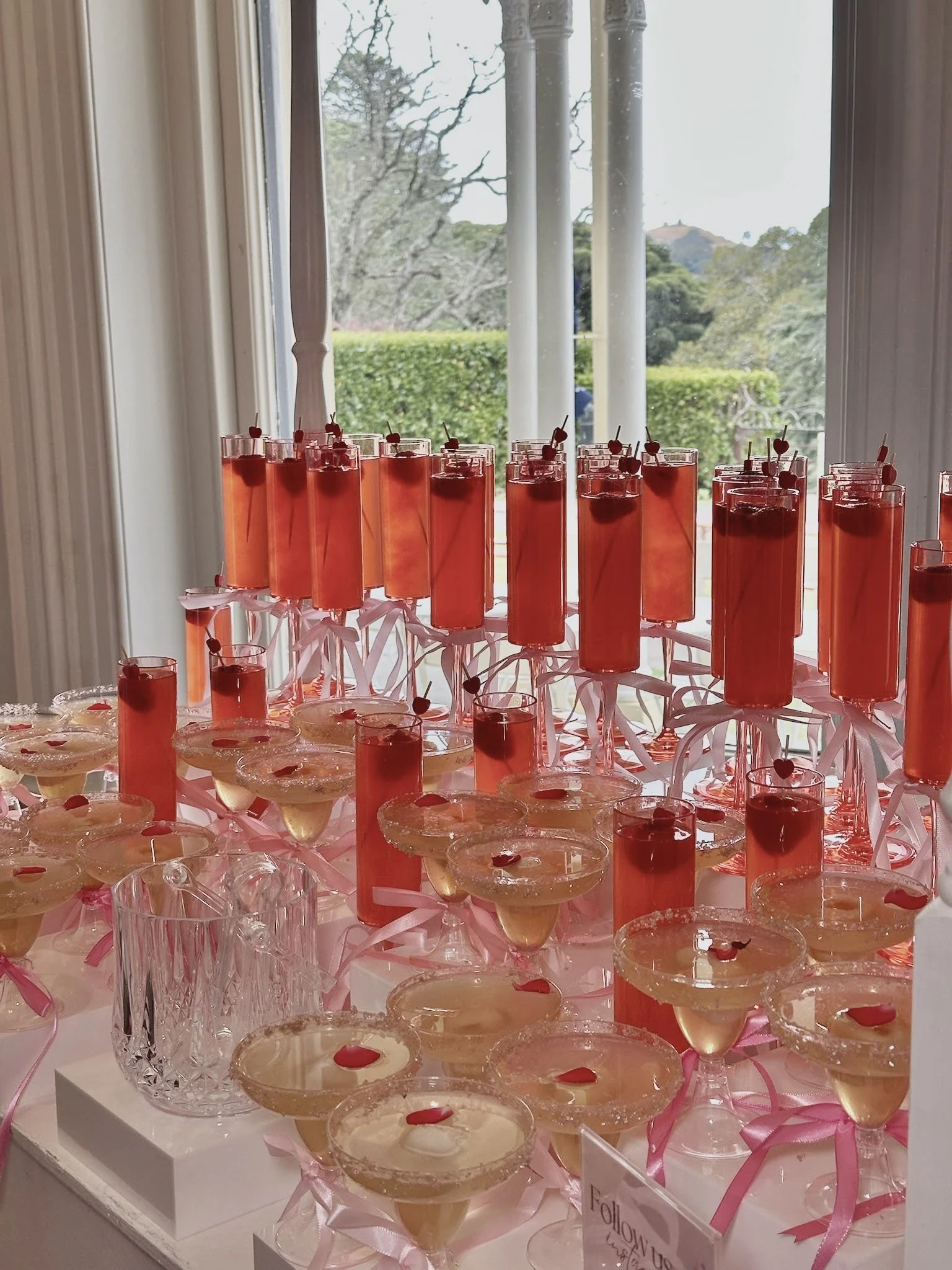 A display of pink-colored beverages in tall glasses with small heart-topped skewers, some with cherries, set on a table with pink ribbons and cake slices, near a window with outdoors visible.