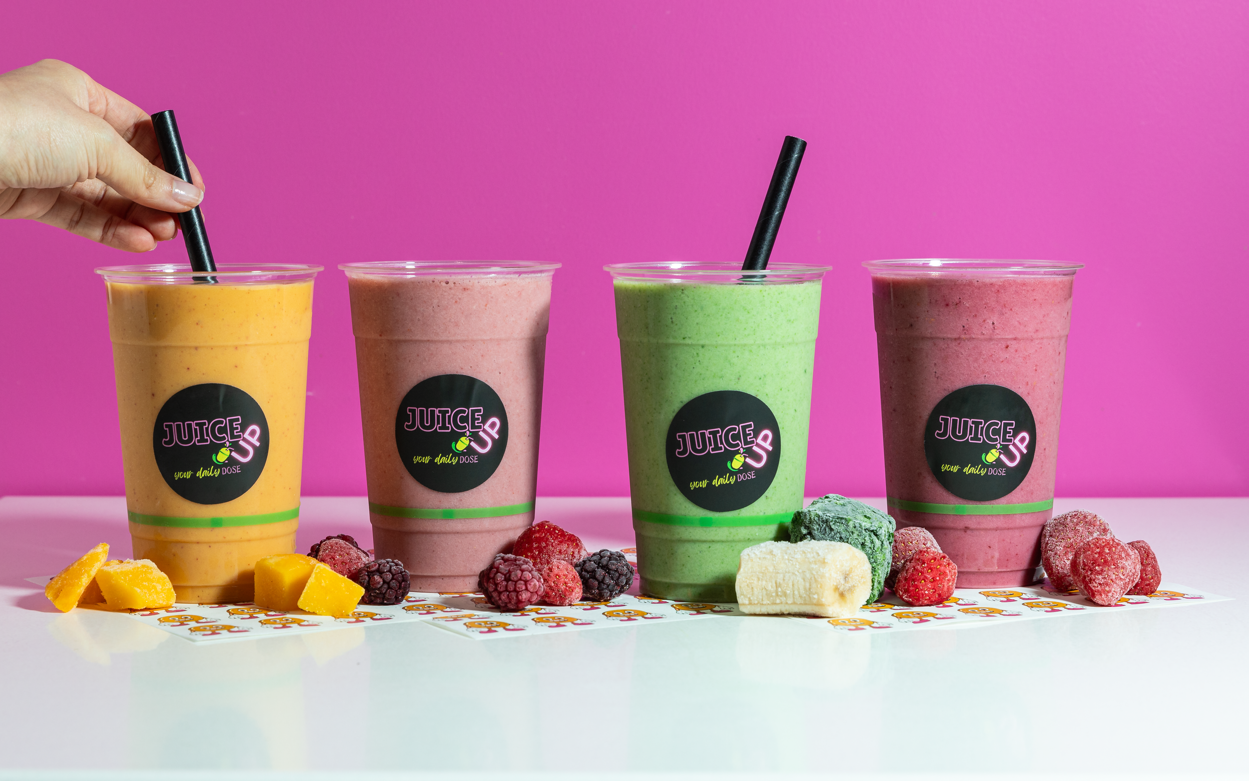 Four colorful smoothies in clear plastic cups with black straws, placed on a white surface with scattered fruit candies and a paper with a logo, against a pink background.