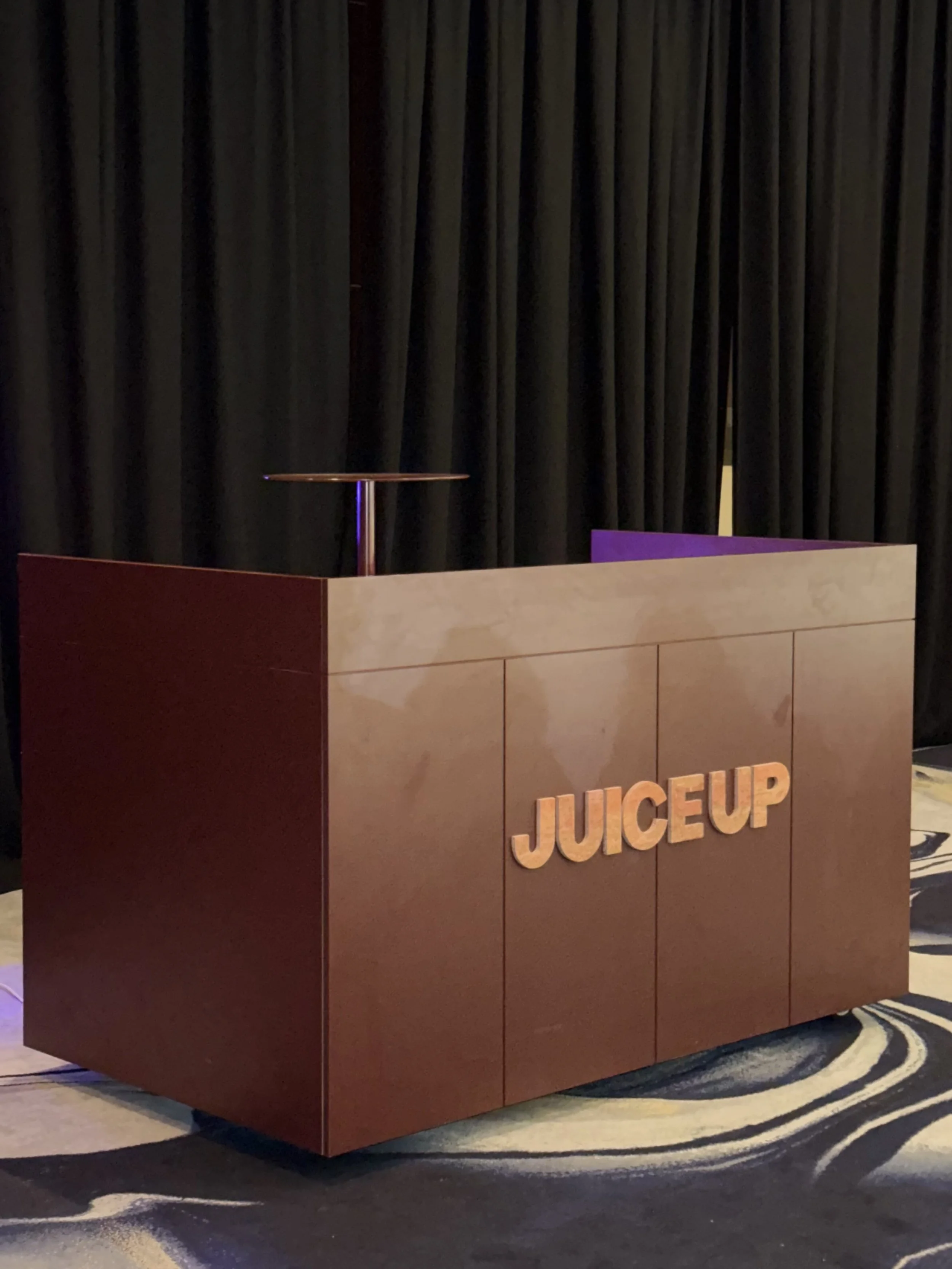A red and beige juice bar counter with the word 'JUICEUP' in wooden letters on the front, set against a dark curtain backdrop in a carpeted conference or event space.