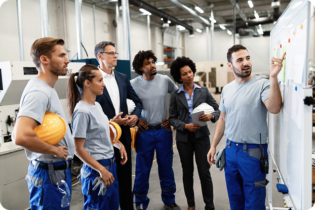 Group of factory workers and a supervisor in an industrial setting, discussing safety plans on a whiteboard.