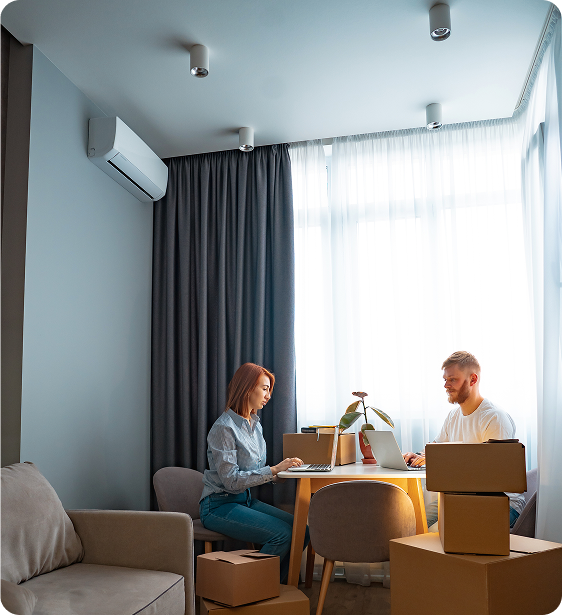 A woman and a man working on laptops at a dining table in a bright living room with large windows, curtains, and an air conditioning unit on the wall.